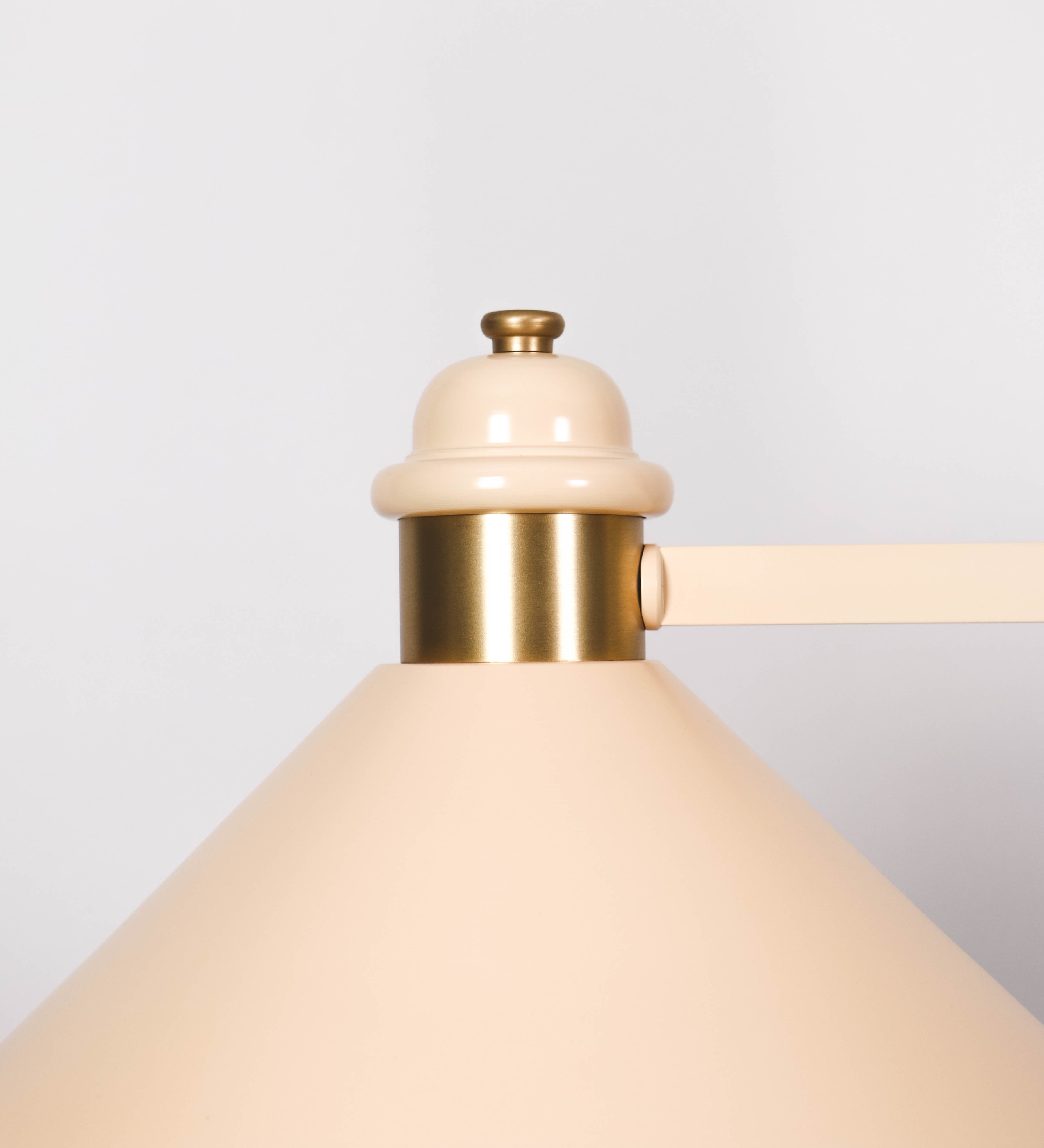 Suspensão de teto moderna e sofisticada. A suspensão é composta por uma base retangular em latão escovado, de onde desce um tubo cilíndrico até a estrutura principal e permite ajustar a altura da suspensão. A estrutura principal é composta por uma barra horizontal bege clara, que sustenta nas extremidades dois chapéus cónicos invertidos no mesmo tom, com interior em latão escovado. No centro da barra horizontal está uma pequena placa oval, em latão escovado, com o ano 1930 inscrito em relevo.