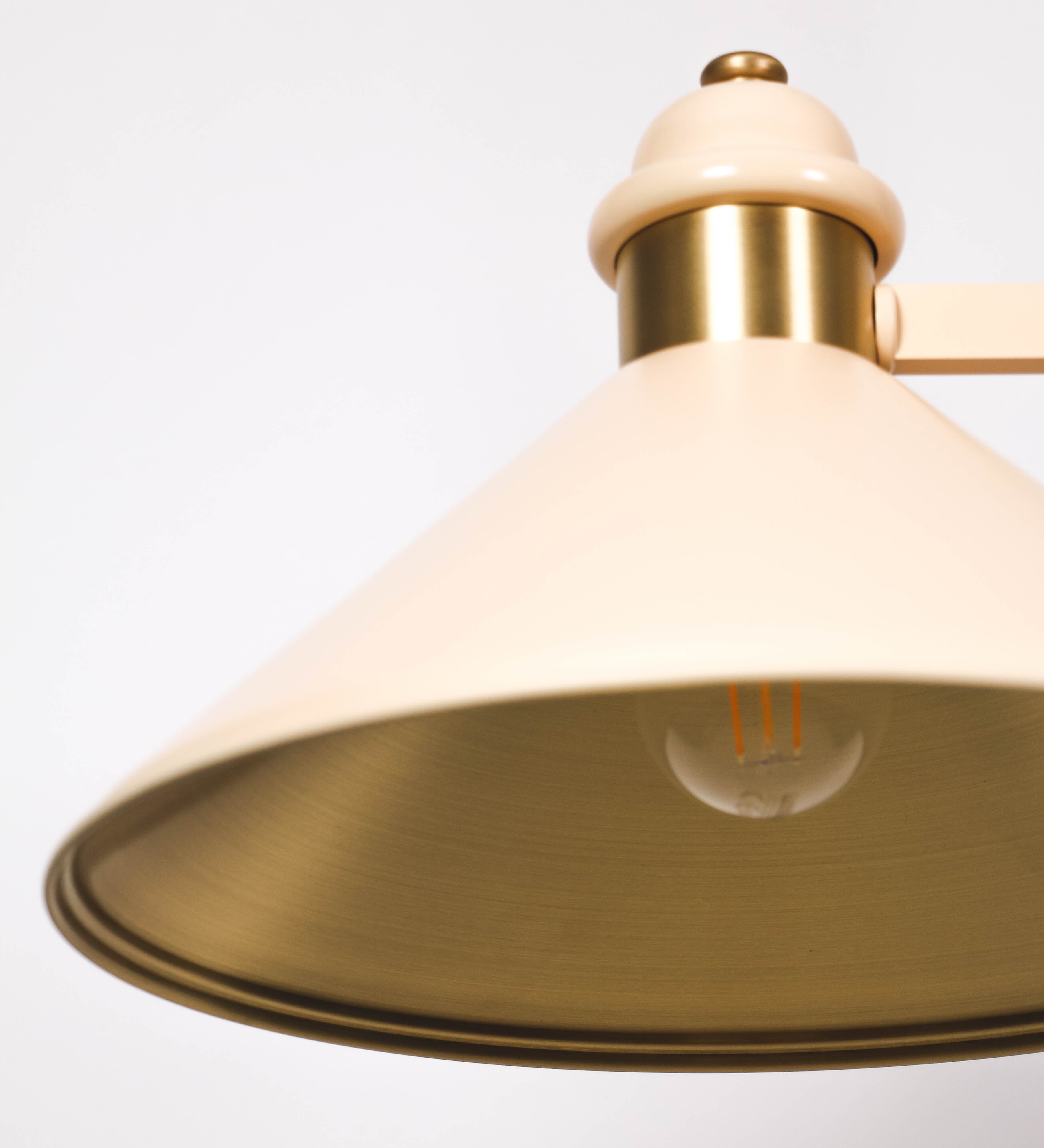 Suspensão de teto moderna e sofisticada. A suspensão é composta por uma base retangular em latão escovado, de onde desce um tubo cilíndrico até a estrutura principal e permite ajustar a altura da suspensão. A estrutura principal é composta por uma barra horizontal bege clara, que sustenta nas extremidades dois chapéus cónicos invertidos no mesmo tom, com interior em latão escovado. No centro da barra horizontal está uma pequena placa oval, em latão escovado, com o ano 1930 inscrito em relevo.