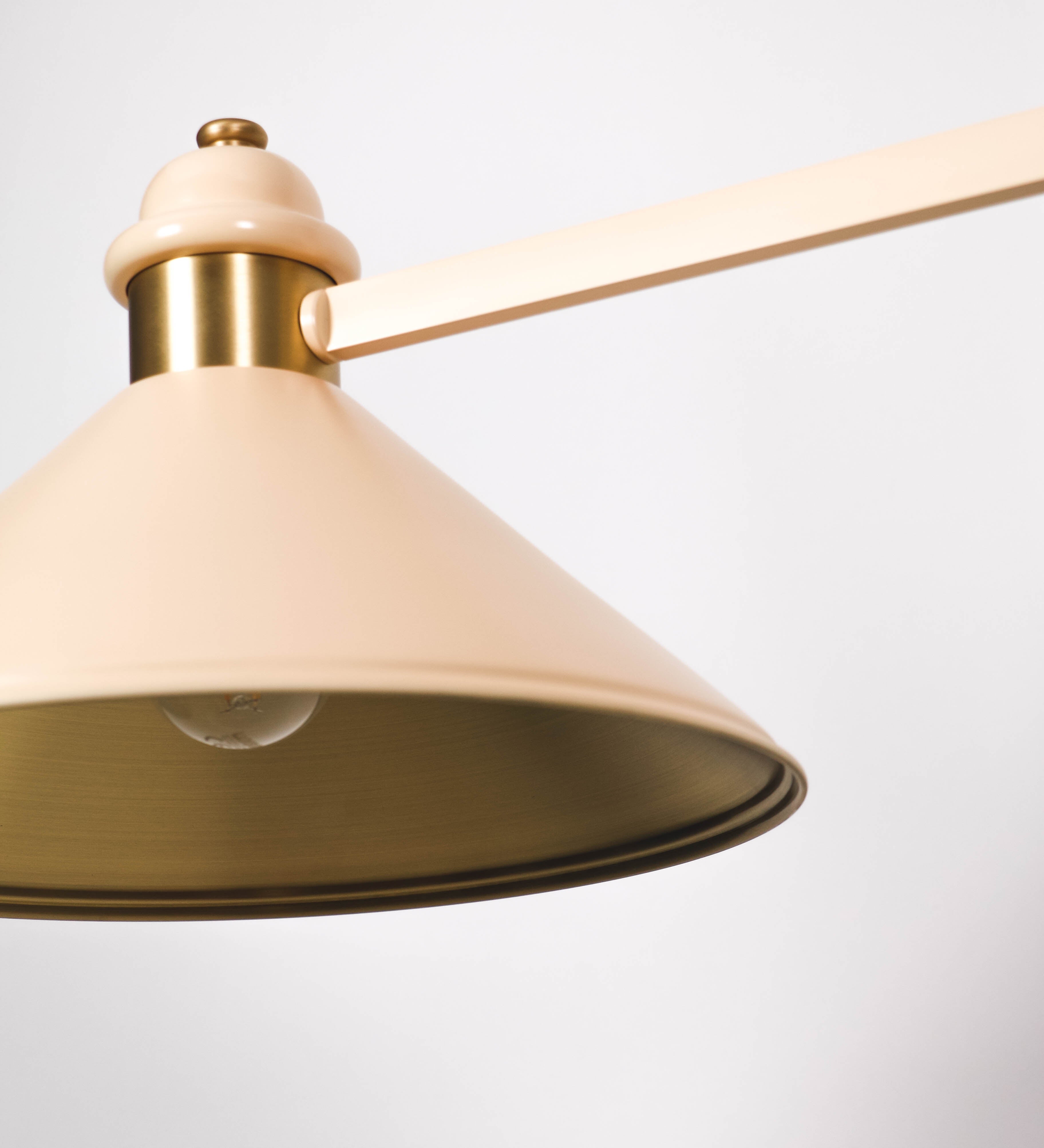 Suspensão de teto moderna e sofisticada. A suspensão é composta por uma base retangular em latão escovado, de onde desce um tubo cilíndrico até a estrutura principal e permite ajustar a altura da suspensão. A estrutura principal é composta por uma barra horizontal bege clara, que sustenta nas extremidades dois chapéus cónicos invertidos no mesmo tom, com interior em latão escovado. No centro da barra horizontal está uma pequena placa oval, em latão escovado, com o ano 1930 inscrito em relevo.