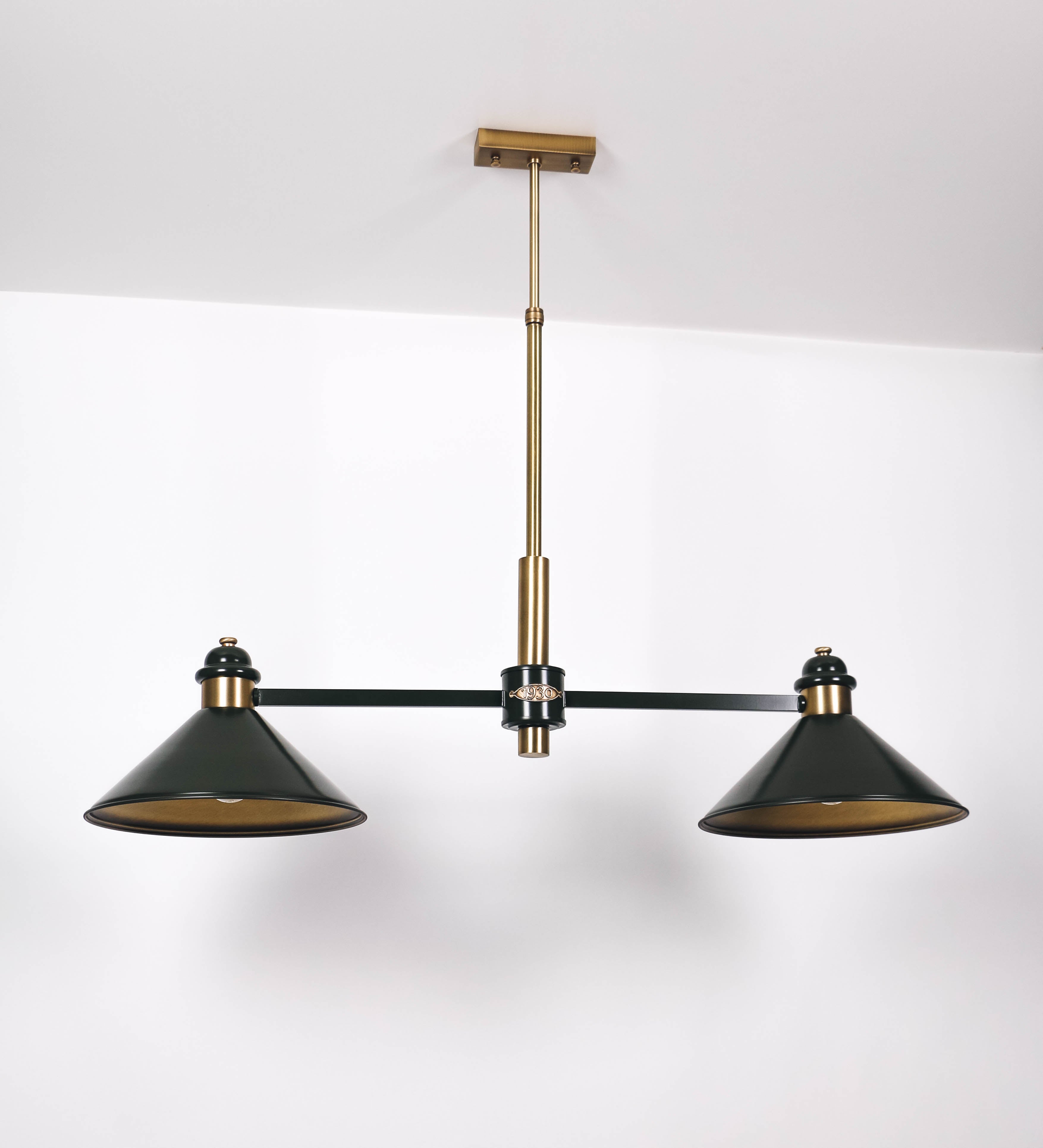 Suspensão de teto moderna e sofisticada. A suspensão é composta por uma base retangular em ouro velho, de onde desce um tubo cilíndrico até a estrutura principal e permite ajustar a altura da suspensão. A estrutura principal é composta por uma barra horizontal verde-azeitona, que sustenta nas extremidades dois chapéus cónicos invertidos no mesmo tom, com interior em ouro velho. No centro da barra horizontal está uma pequena placa oval, em ouro velho, com o ano 1930 inscrito em relevo.