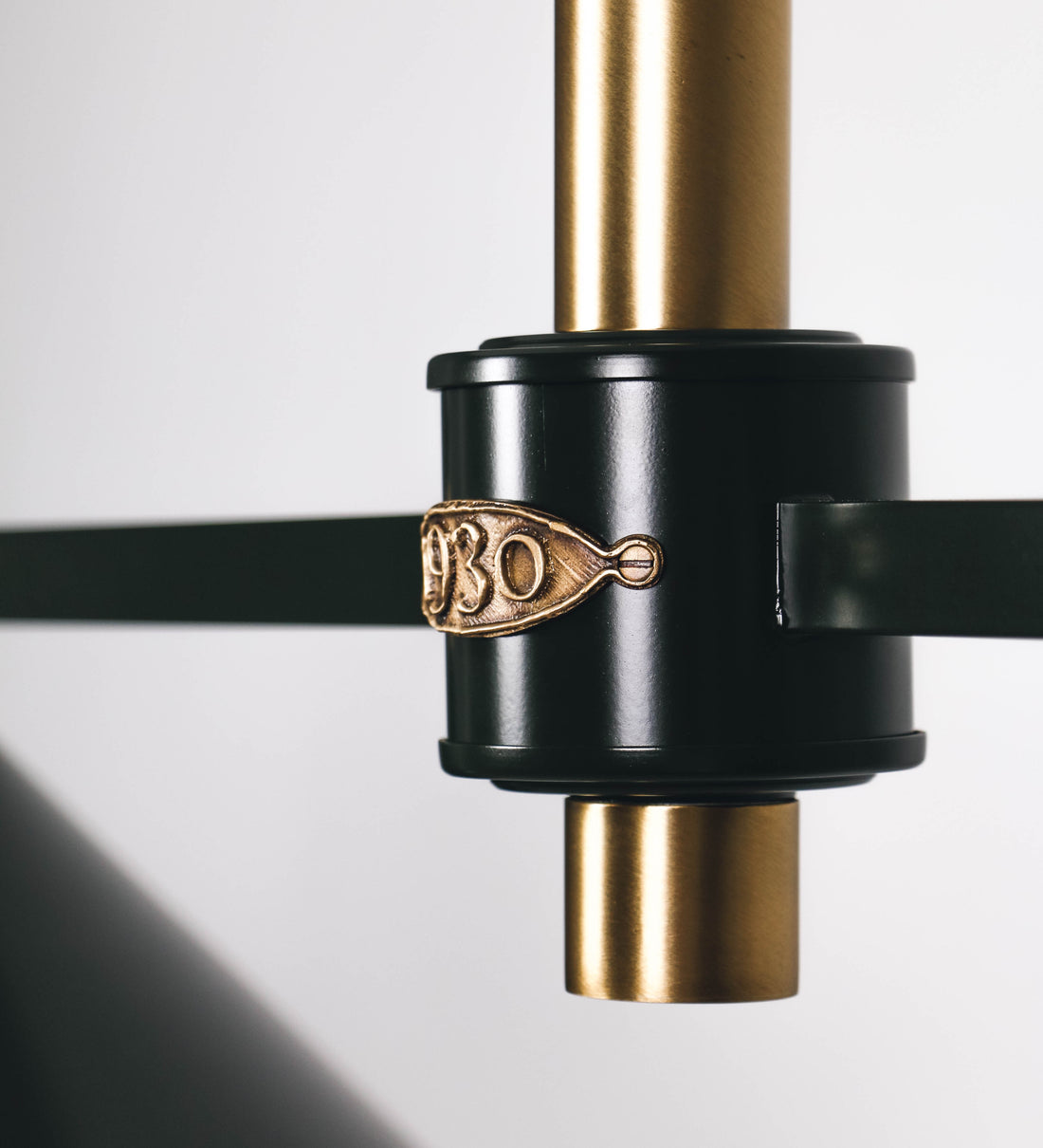 Suspensão de teto moderna e sofisticada. A suspensão é composta por uma base retangular em ouro velho, de onde desce um tubo cilíndrico até a estrutura principal e permite ajustar a altura da suspensão. A estrutura principal é composta por uma barra horizontal verde-azeitona, que sustenta nas extremidades dois chapéus cónicos invertidos no mesmo tom, com interior em ouro velho. No centro da barra horizontal está uma pequena placa oval, em ouro velho, com o ano 1930 inscrito em relevo.