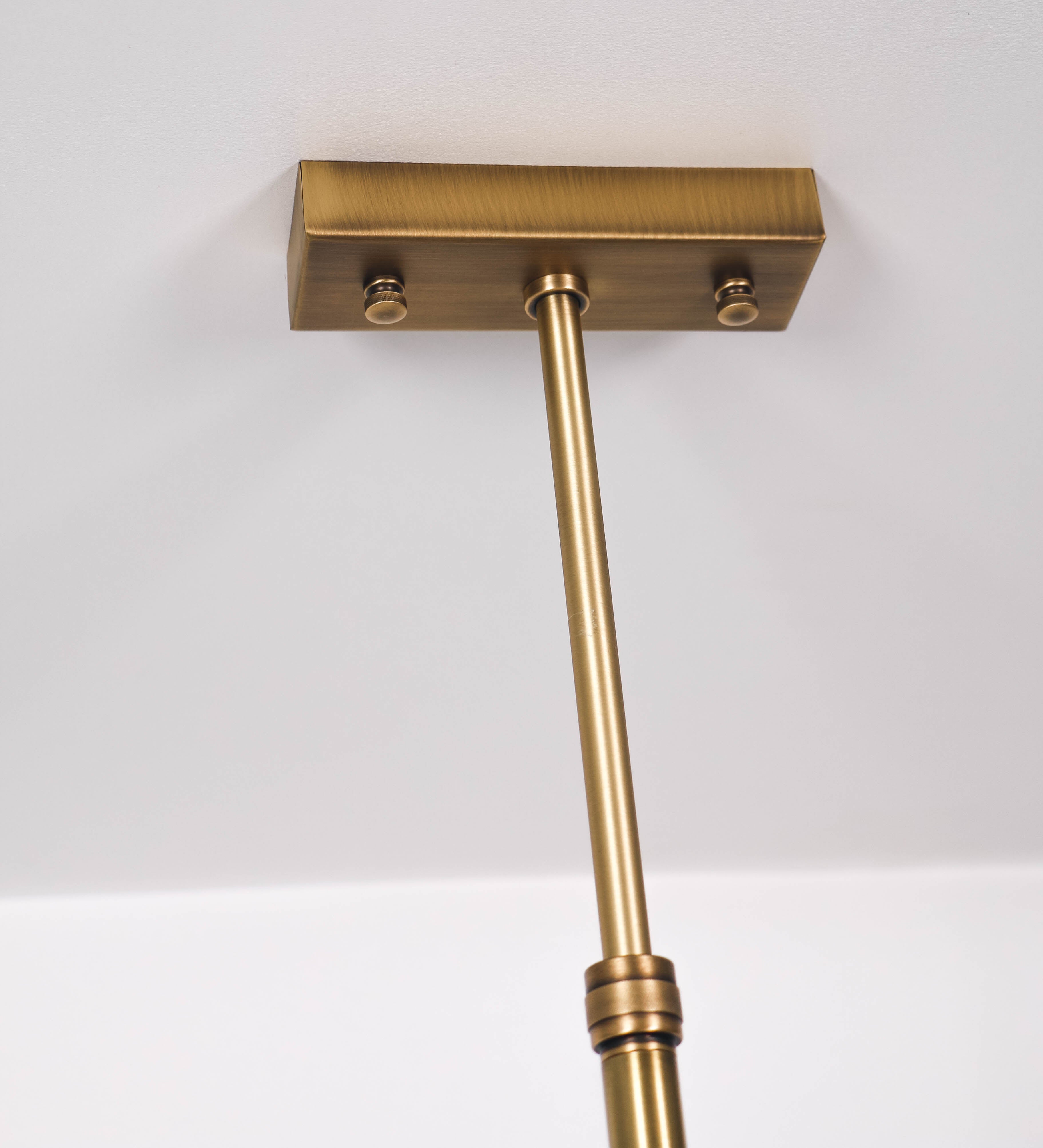 Suspensão de teto moderna e sofisticada. A suspensão é composta por uma base retangular em ouro velho, de onde desce um tubo cilíndrico até a estrutura principal e permite ajustar a altura da suspensão. A estrutura principal é composta por uma barra horizontal verde-azeitona, que sustenta nas extremidades dois chapéus cónicos invertidos no mesmo tom, com interior em ouro velho. No centro da barra horizontal está uma pequena placa oval, em ouro velho, com o ano 1930 inscrito em relevo.