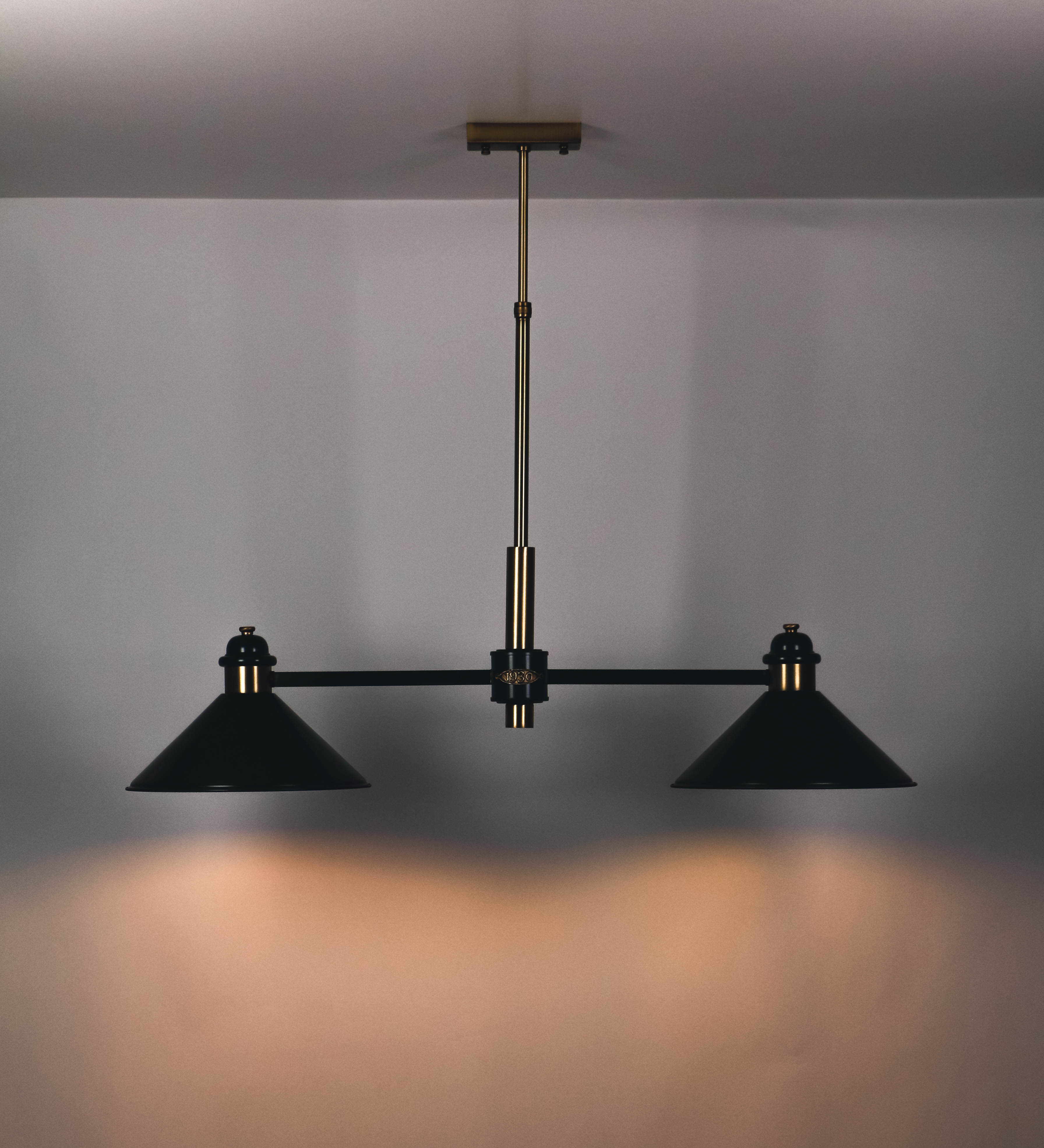 Suspensão de teto moderna e sofisticada. A suspensão é composta por uma base retangular em ouro velho, de onde desce um tubo cilíndrico até a estrutura principal e permite ajustar a altura da suspensão. A estrutura principal é composta por uma barra horizontal verde-azeitona, que sustenta nas extremidades dois chapéus cónicos invertidos no mesmo tom, com interior em ouro velho. No centro da barra horizontal está uma pequena placa oval, em ouro velho, com o ano 1930 inscrito em relevo.