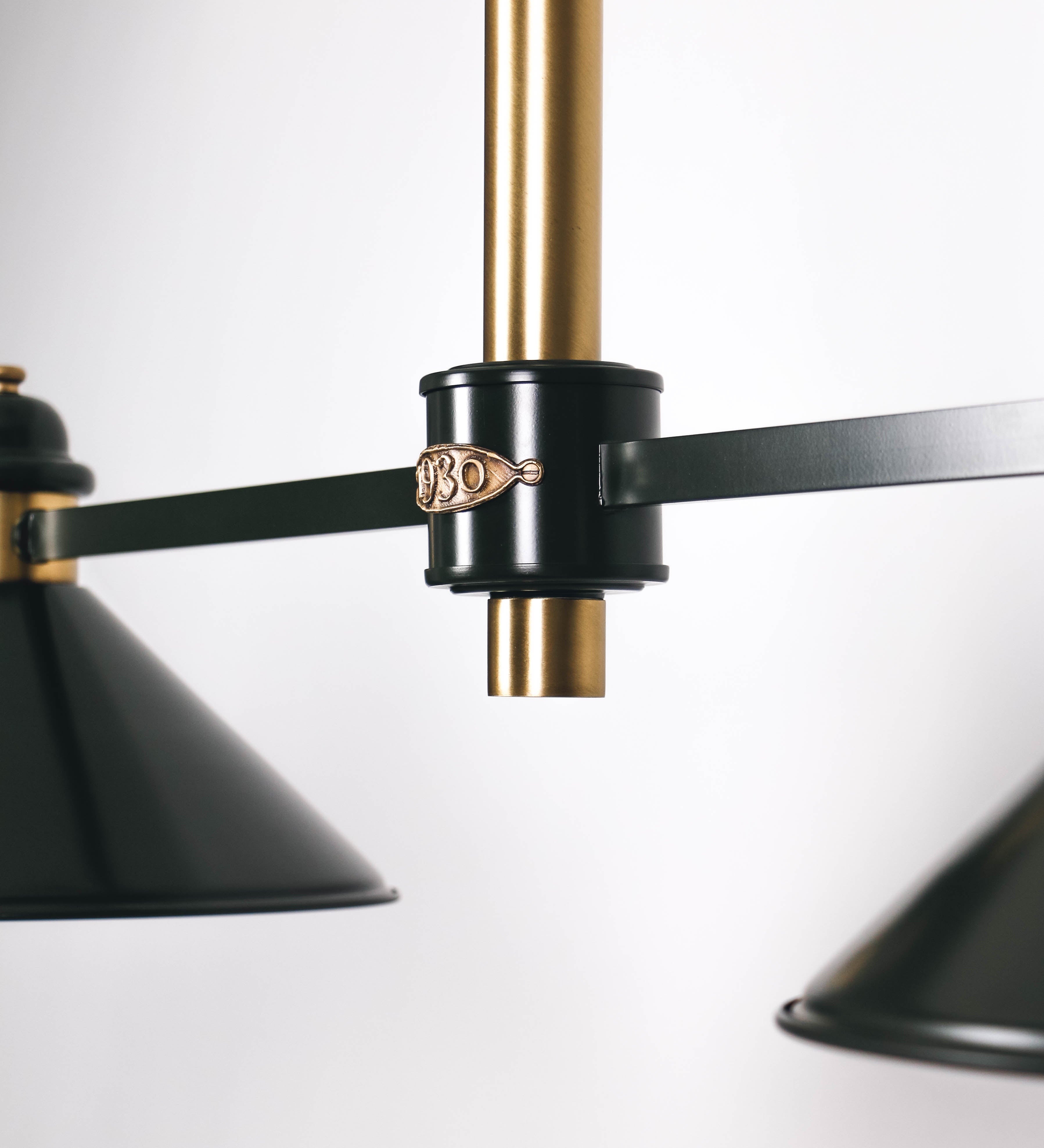 Suspensão de teto moderna e sofisticada. A suspensão é composta por uma base retangular em ouro velho, de onde desce um tubo cilíndrico até a estrutura principal e permite ajustar a altura da suspensão. A estrutura principal é composta por uma barra horizontal verde-azeitona, que sustenta nas extremidades dois chapéus cónicos invertidos no mesmo tom, com interior em ouro velho. No centro da barra horizontal está uma pequena placa oval, em ouro velho, com o ano 1930 inscrito em relevo.