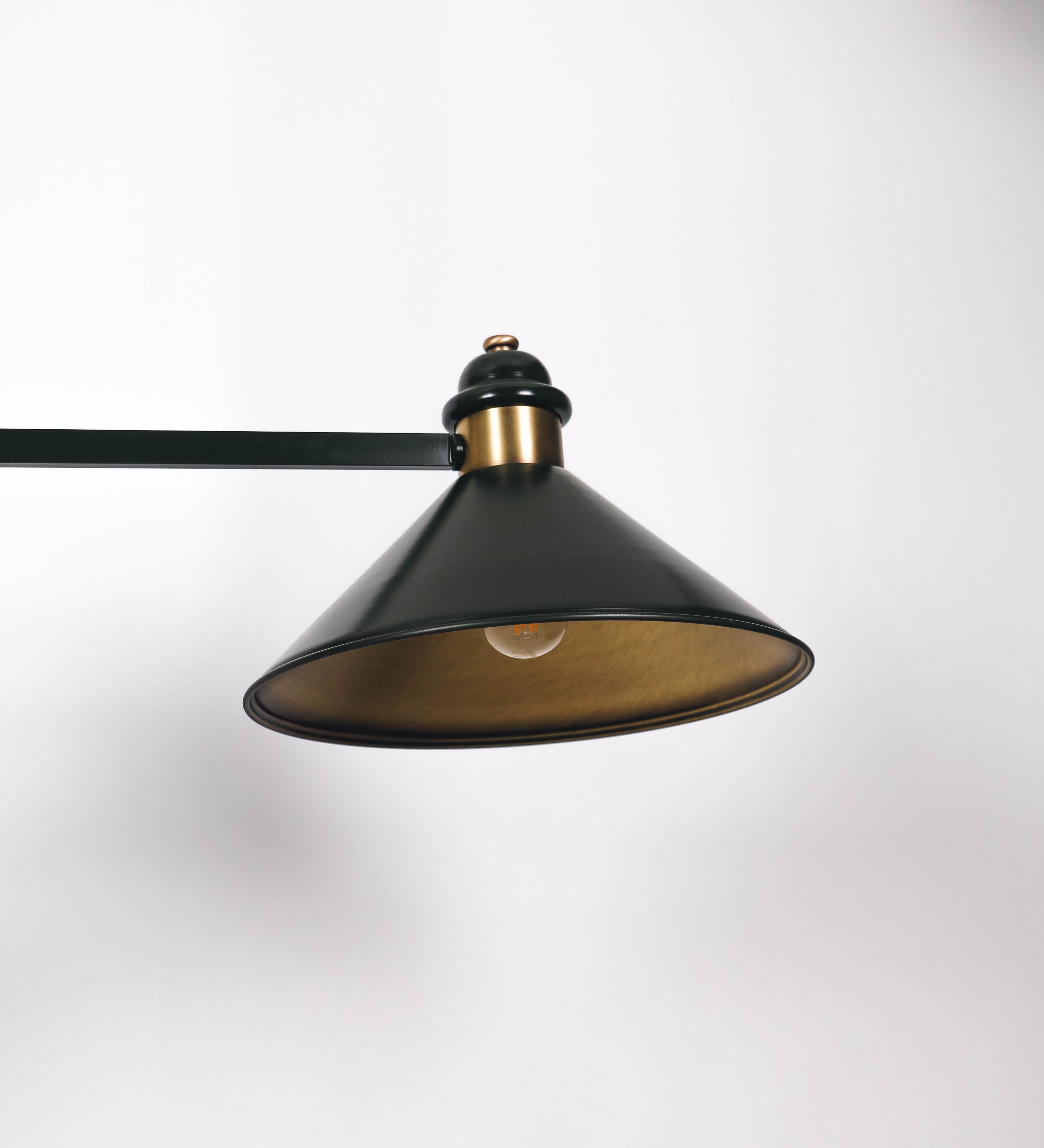 Suspensão de teto moderna e sofisticada. A suspensão é composta por uma base retangular em ouro velho, de onde desce um tubo cilíndrico até a estrutura principal e permite ajustar a altura da suspensão. A estrutura principal é composta por uma barra horizontal verde-azeitona, que sustenta nas extremidades dois chapéus cónicos invertidos no mesmo tom, com interior em ouro velho. No centro da barra horizontal está uma pequena placa oval, em ouro velho, com o ano 1930 inscrito em relevo.