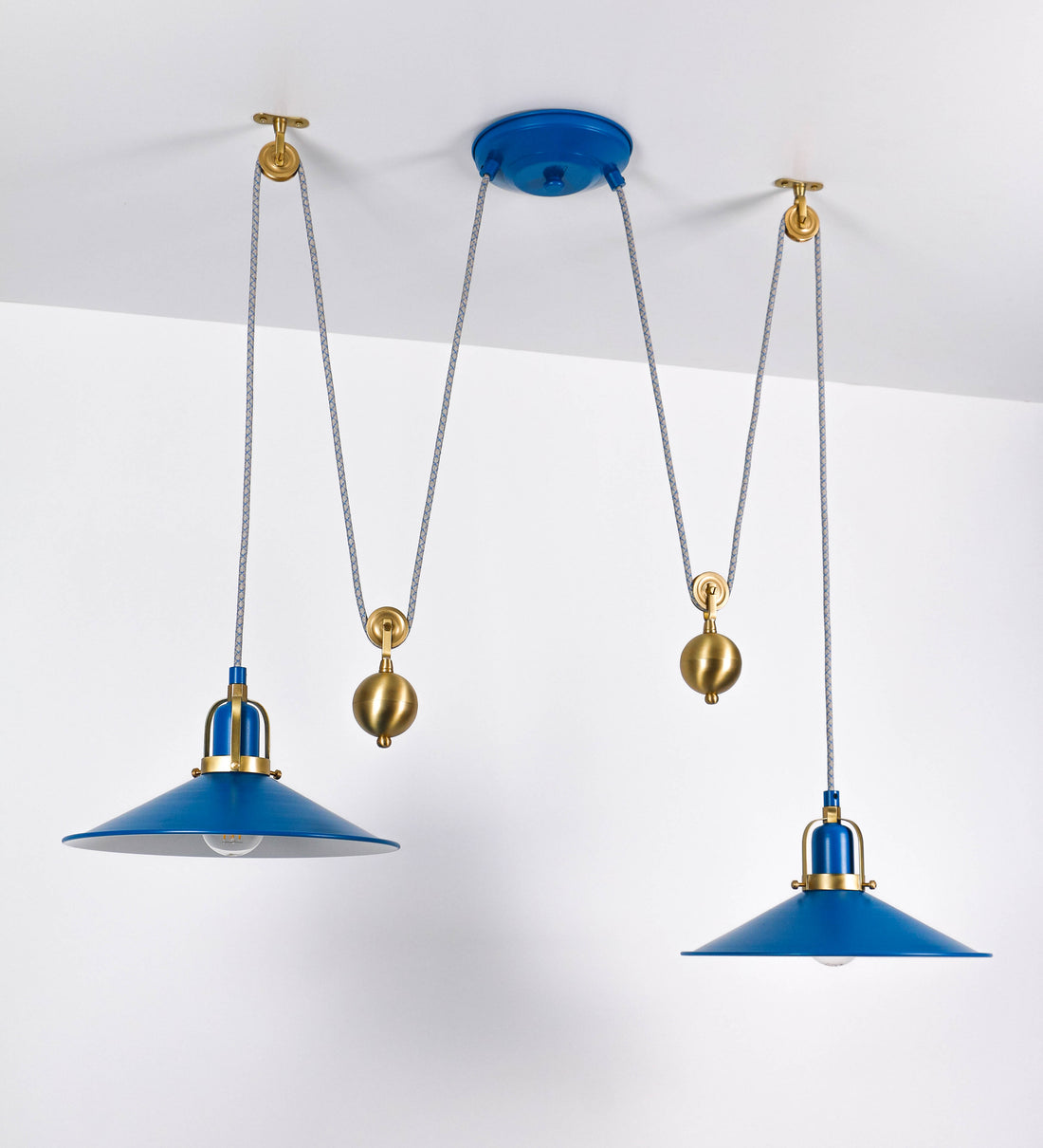 Suspensão de teto dupla composta por uma base circular azul vintage de onde descem dois cabos revestidos têxtil com linhas azuis e beges. Cada cabo passa por uma roldana em latão escovado fixa ao teto e desce até aos chapéus. Entre a base e as roldanas cada cabo sustenta uma esfera em latão escovado que atua como contrapeso para ajustar a altura. Os dois chapéus são azuis de formato cónico invertido e interior branco. No topo de cada chapéu há um suporte em latão escovado que conecta os chapéus aos cabos.