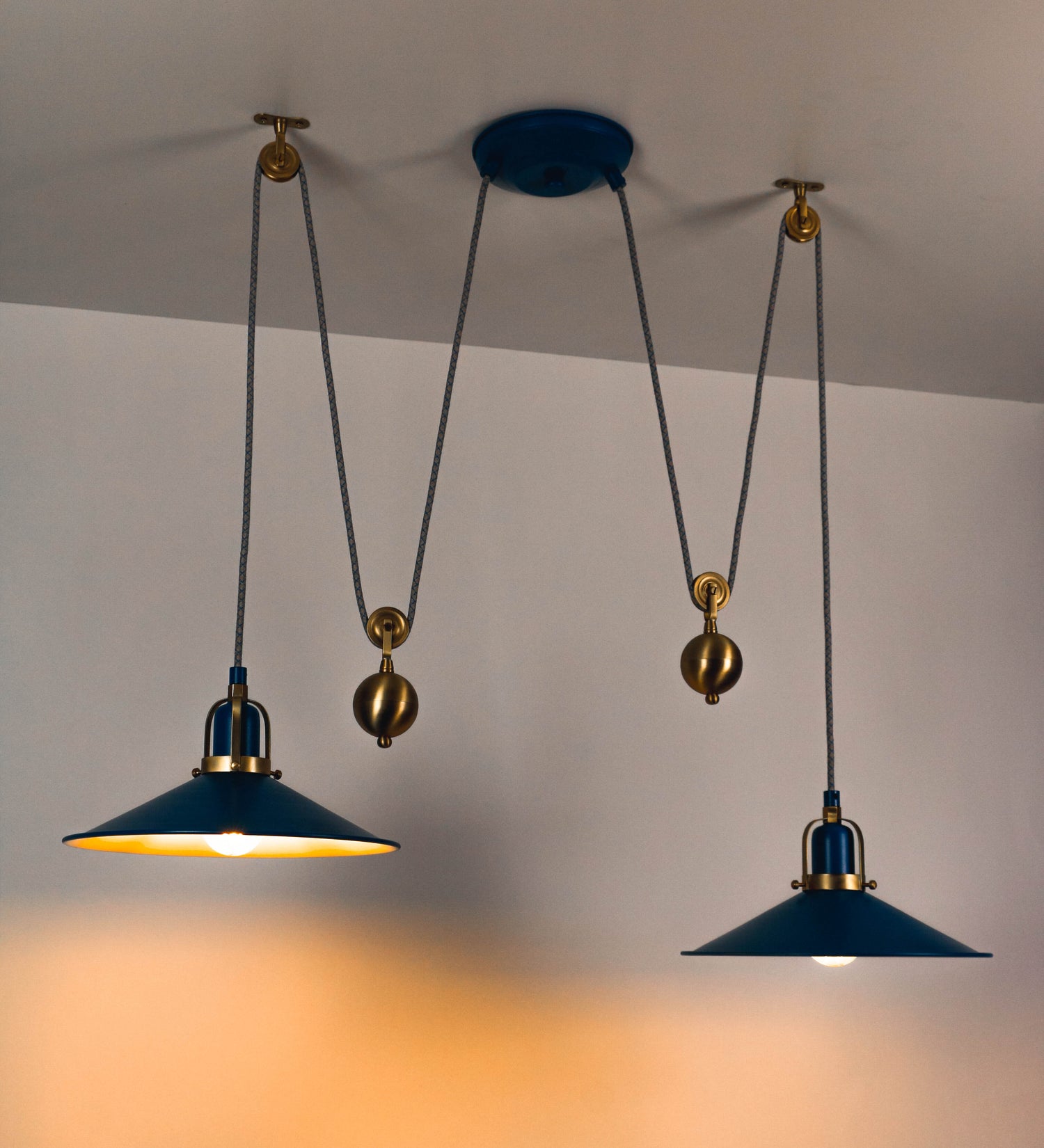 Suspensão de teto dupla composta por uma base circular azul vintage de onde descem dois cabos revestidos têxtil com linhas azuis e beges. Cada cabo passa por uma roldana em latão escovado fixa ao teto e desce até aos chapéus. Entre a base e as roldanas cada cabo sustenta uma esfera em latão escovado que atua como contrapeso para ajustar a altura. Os dois chapéus são azuis de formato cónico invertido e interior branco. No topo de cada chapéu há um suporte em latão escovado que conecta os chapéus aos cabos.