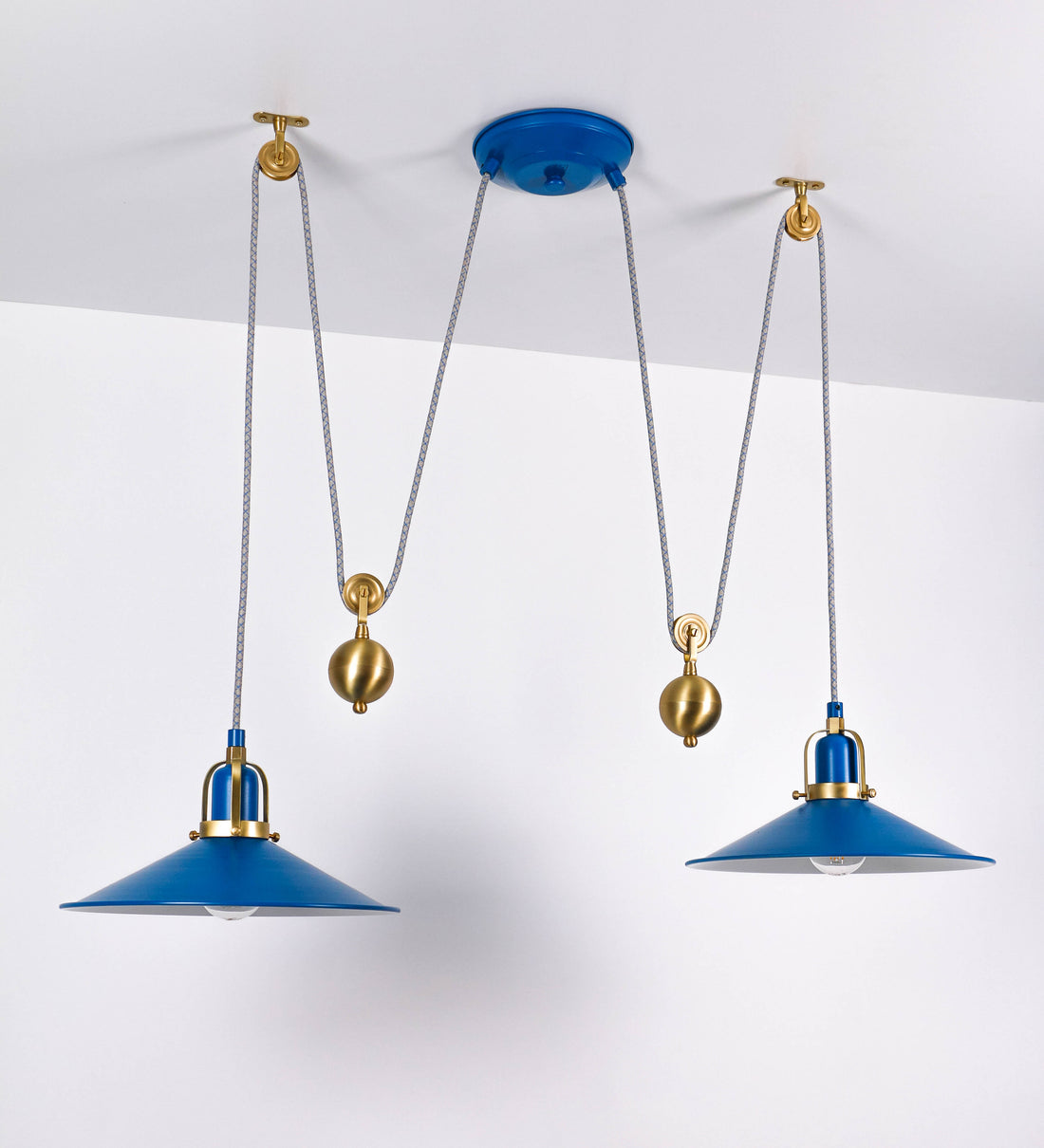 Suspensão de teto dupla composta por uma base circular azul vintage de onde descem dois cabos revestidos têxtil com linhas azuis e beges. Cada cabo passa por uma roldana em latão escovado fixa ao teto e desce até aos chapéus. Entre a base e as roldanas cada cabo sustenta uma esfera em latão escovado que atua como contrapeso para ajustar a altura. Os dois chapéus são azuis de formato cónico invertido e interior branco. No topo de cada chapéu há um suporte em latão escovado que conecta os chapéus aos cabos.
