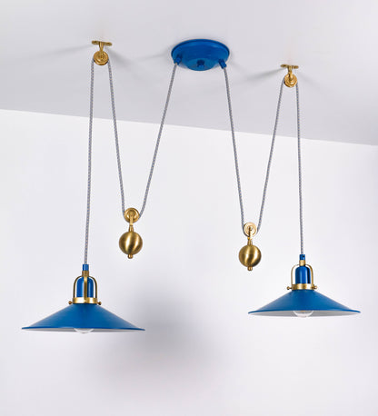 Suspensão de teto dupla composta por uma base circular azul vintage de onde descem dois cabos revestidos têxtil com linhas azuis e beges. Cada cabo passa por uma roldana em latão escovado fixa ao teto e desce até aos chapéus. Entre a base e as roldanas cada cabo sustenta uma esfera em latão escovado que atua como contrapeso para ajustar a altura. Os dois chapéus são azuis de formato cónico invertido e interior branco. No topo de cada chapéu há um suporte em latão escovado que conecta os chapéus aos cabos.