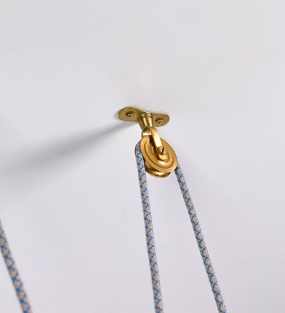 Suspensão de teto dupla composta por uma base circular azul vintage de onde descem dois cabos revestidos têxtil com linhas azuis e beges. Cada cabo passa por uma roldana em latão escovado fixa ao teto e desce até aos chapéus. Entre a base e as roldanas cada cabo sustenta uma esfera em latão escovado que atua como contrapeso para ajustar a altura. Os dois chapéus são azuis de formato cónico invertido e interior branco. No topo de cada chapéu há um suporte em latão escovado que conecta os chapéus aos cabos.