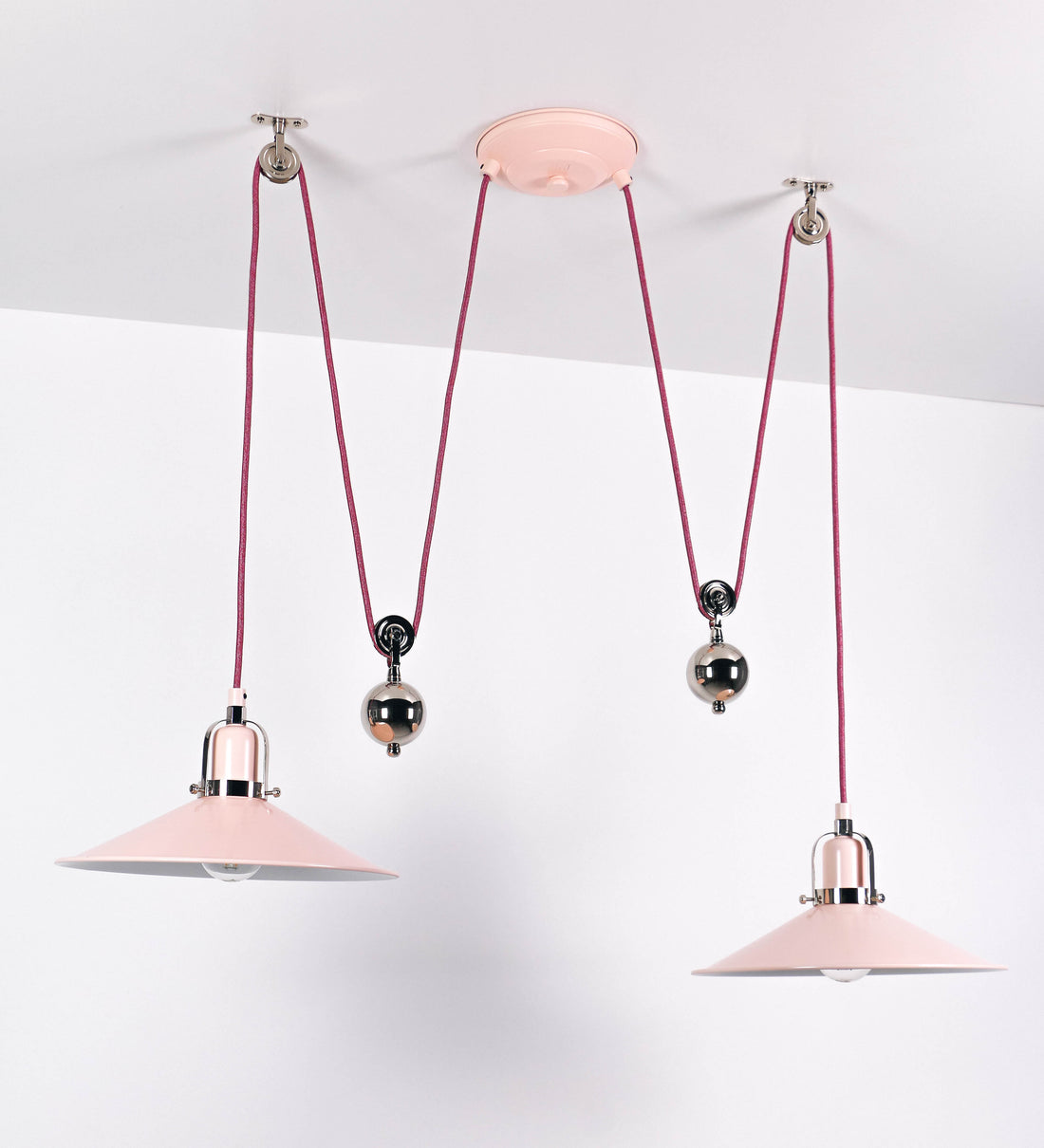 Suspensão de teto dupla composta por uma base circular rosa clara, de onde descem dois cabos revestidos têxtil rosa velho. Cada cabo passa por uma roldana, em níquel polido fixa ao teto, e desce até aos chapéus. Entre a base e as roldanas, cada cabo sustenta uma esfera em níquel polido, que atua como contrapeso para ajustar a altura. Os dois chapéus são rosa claro, de formato cónico invertido e interior branco. No topo de cada chapéu há um suporte em níquel polido que conecta os chapéus aos cabos.