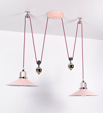 Suspensão de teto dupla composta por uma base circular rosa clara, de onde descem dois cabos revestidos têxtil rosa velho. Cada cabo passa por uma roldana, em níquel polido fixa ao teto, e desce até aos chapéus. Entre a base e as roldanas, cada cabo sustenta uma esfera em níquel polido, que atua como contrapeso para ajustar a altura. Os dois chapéus são rosa claro, de formato cónico invertido e interior branco. No topo de cada chapéu há um suporte em níquel polido que conecta os chapéus aos cabos.