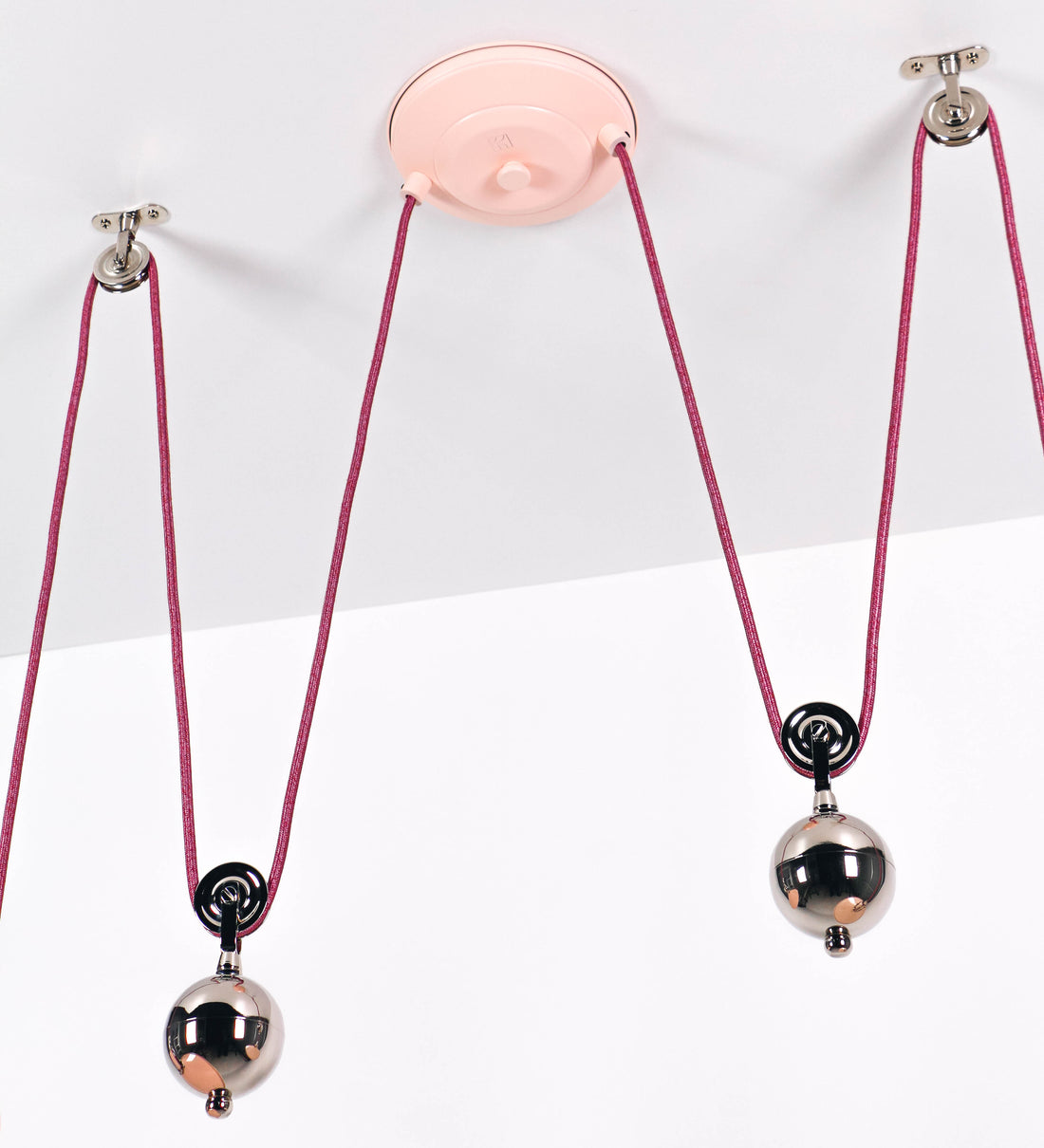 Suspensão de teto dupla composta por uma base circular rosa clara, de onde descem dois cabos revestidos têxtil rosa velho. Cada cabo passa por uma roldana, em níquel polido fixa ao teto, e desce até aos chapéus. Entre a base e as roldanas, cada cabo sustenta uma esfera em níquel polido, que atua como contrapeso para ajustar a altura. Os dois chapéus são rosa claro, de formato cónico invertido e interior branco. No topo de cada chapéu há um suporte em níquel polido que conecta os chapéus aos cabos.