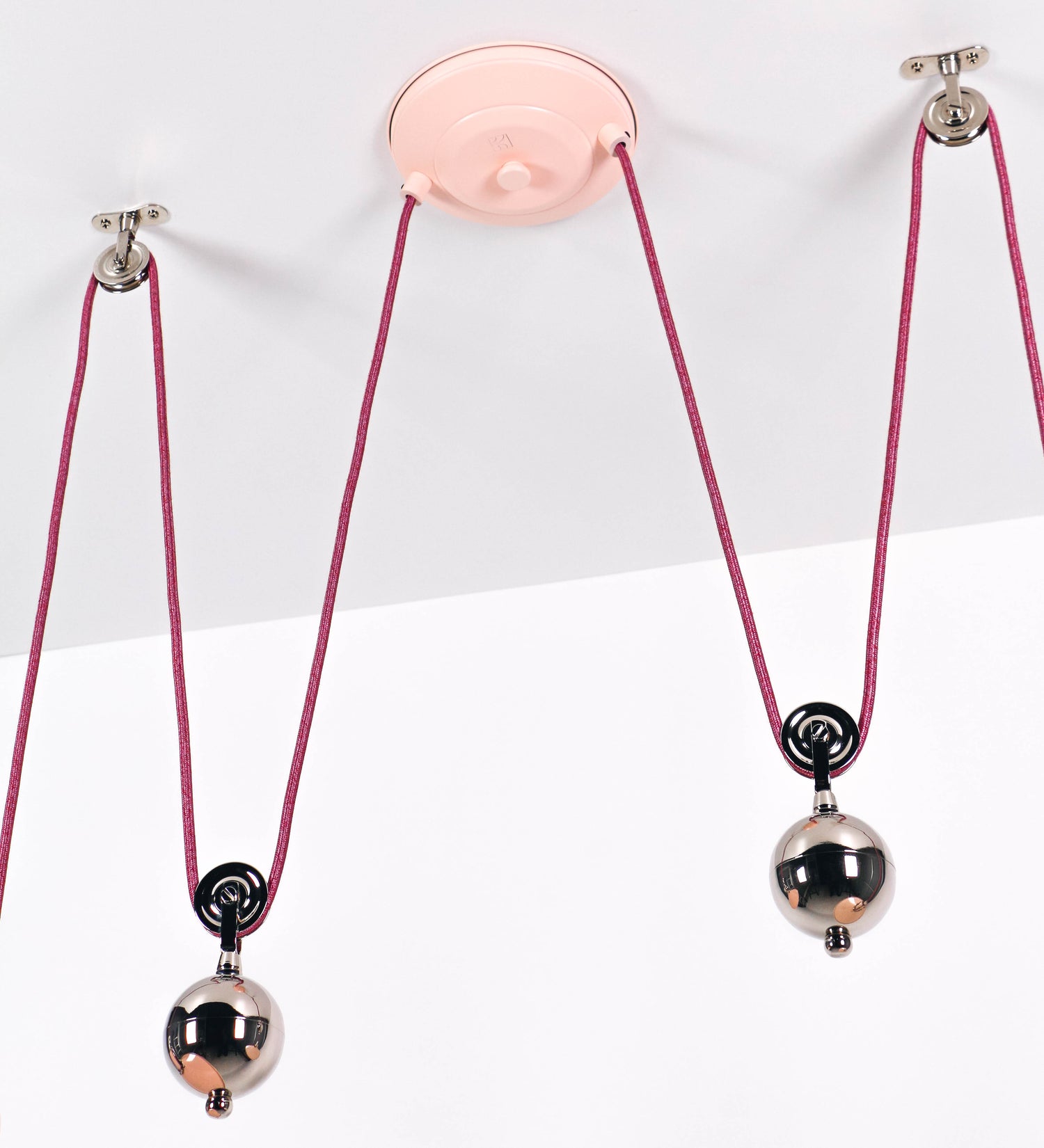 Suspensão de teto dupla composta por uma base circular rosa clara, de onde descem dois cabos revestidos têxtil rosa velho. Cada cabo passa por uma roldana, em níquel polido fixa ao teto, e desce até aos chapéus. Entre a base e as roldanas, cada cabo sustenta uma esfera em níquel polido, que atua como contrapeso para ajustar a altura. Os dois chapéus são rosa claro, de formato cónico invertido e interior branco. No topo de cada chapéu há um suporte em níquel polido que conecta os chapéus aos cabos.
