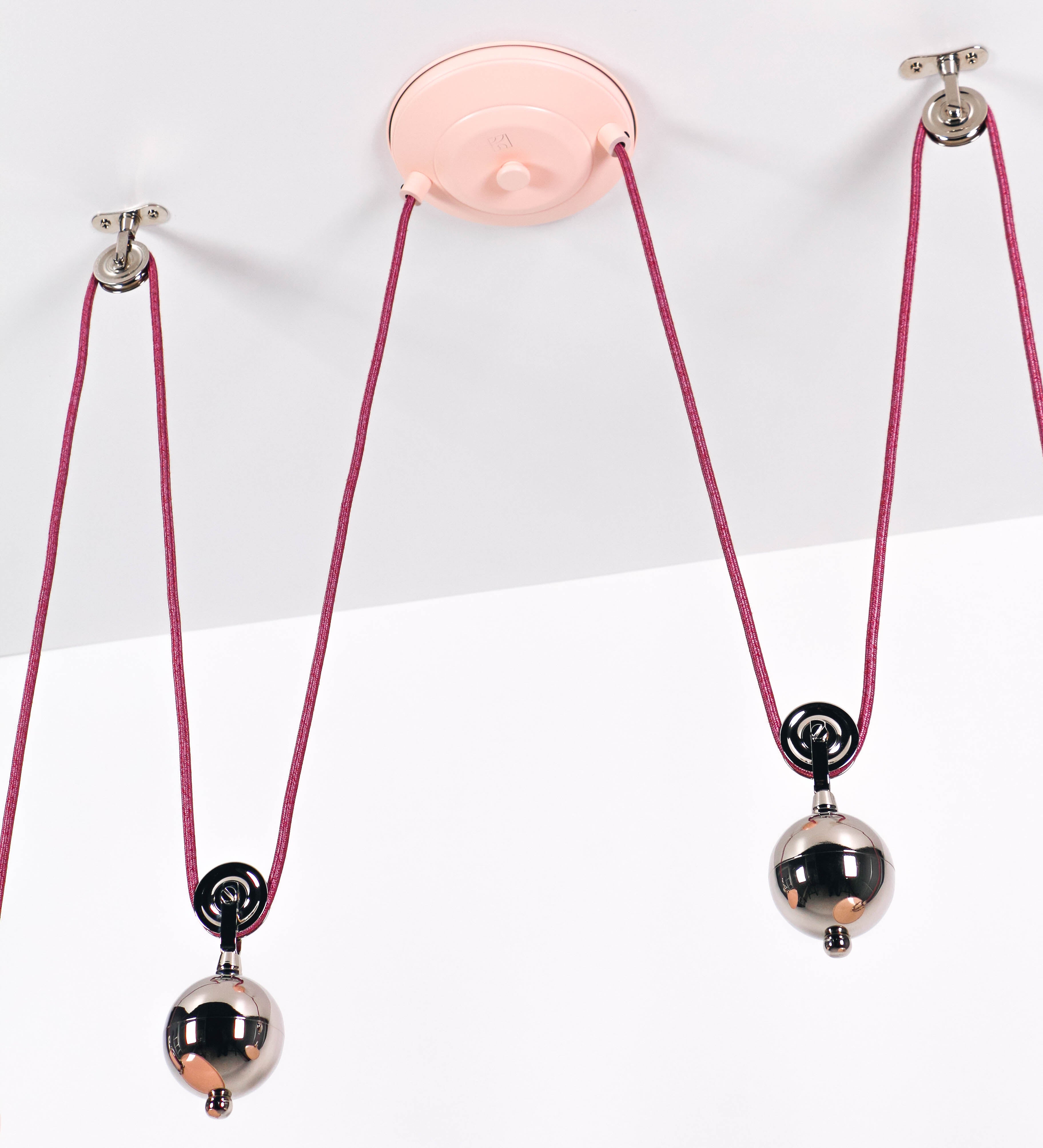 Suspensão de teto dupla composta por uma base circular rosa clara, de onde descem dois cabos revestidos têxtil rosa velho. Cada cabo passa por uma roldana, em níquel polido fixa ao teto, e desce até aos chapéus. Entre a base e as roldanas, cada cabo sustenta uma esfera em níquel polido, que atua como contrapeso para ajustar a altura. Os dois chapéus são rosa claro, de formato cónico invertido e interior branco. No topo de cada chapéu há um suporte em níquel polido que conecta os chapéus aos cabos.