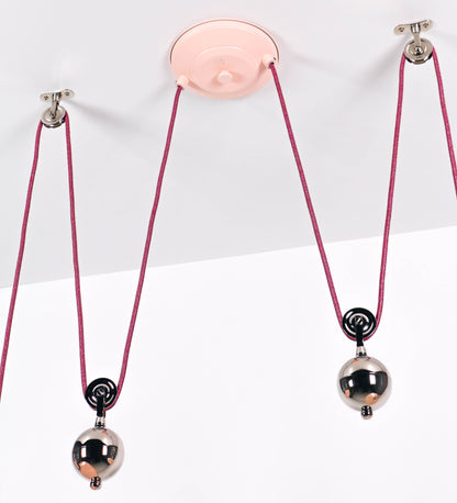 Suspensão de teto dupla composta por uma base circular rosa clara, de onde descem dois cabos revestidos têxtil rosa velho. Cada cabo passa por uma roldana, em níquel polido fixa ao teto, e desce até aos chapéus. Entre a base e as roldanas, cada cabo sustenta uma esfera em níquel polido, que atua como contrapeso para ajustar a altura. Os dois chapéus são rosa claro, de formato cónico invertido e interior branco. No topo de cada chapéu há um suporte em níquel polido que conecta os chapéus aos cabos.