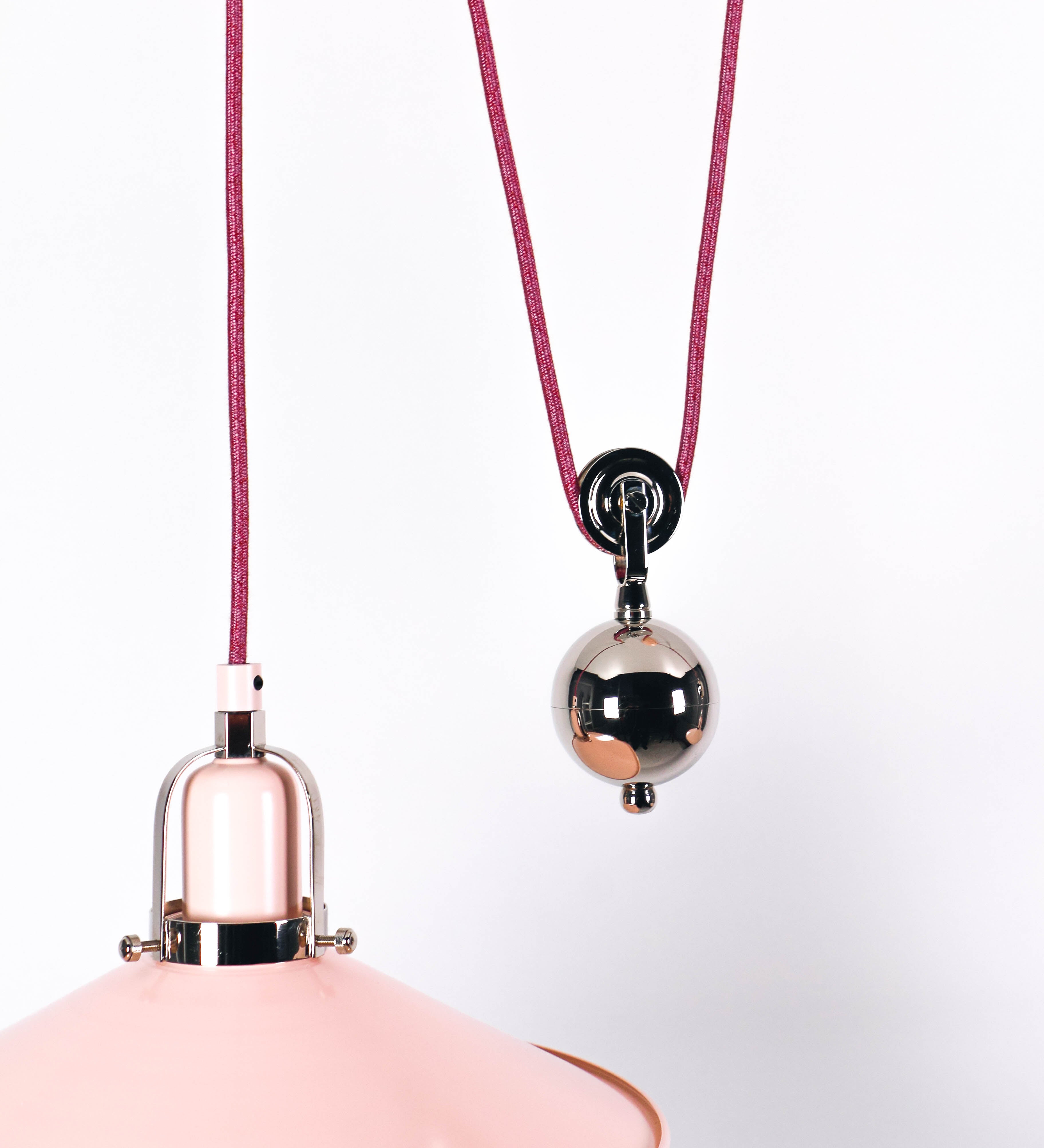 Suspensão de teto dupla composta por uma base circular rosa clara, de onde descem dois cabos revestidos têxtil rosa velho. Cada cabo passa por uma roldana, em níquel polido fixa ao teto, e desce até aos chapéus. Entre a base e as roldanas, cada cabo sustenta uma esfera em níquel polido, que atua como contrapeso para ajustar a altura. Os dois chapéus são rosa claro, de formato cónico invertido e interior branco. No topo de cada chapéu há um suporte em níquel polido que conecta os chapéus aos cabos.