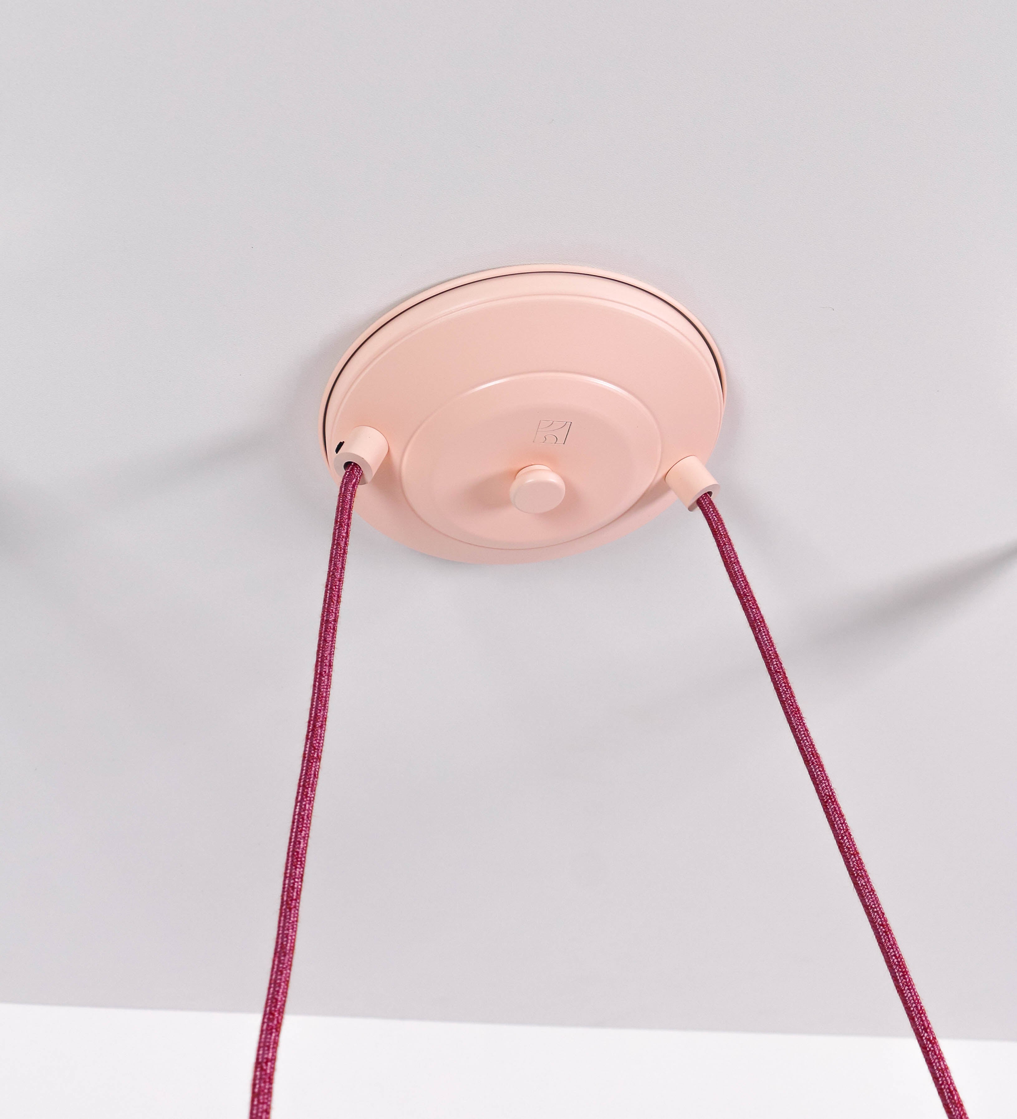 Suspensão de teto dupla composta por uma base circular rosa clara, de onde descem dois cabos revestidos têxtil rosa velho. Cada cabo passa por uma roldana, em níquel polido fixa ao teto, e desce até aos chapéus. Entre a base e as roldanas, cada cabo sustenta uma esfera em níquel polido, que atua como contrapeso para ajustar a altura. Os dois chapéus são rosa claro, de formato cónico invertido e interior branco. No topo de cada chapéu há um suporte em níquel polido que conecta os chapéus aos cabos.