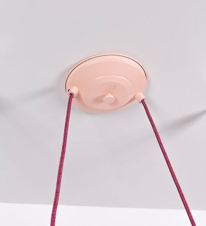 Suspensão de teto dupla composta por uma base circular rosa clara, de onde descem dois cabos revestidos têxtil rosa velho. Cada cabo passa por uma roldana, em níquel polido fixa ao teto, e desce até aos chapéus. Entre a base e as roldanas, cada cabo sustenta uma esfera em níquel polido, que atua como contrapeso para ajustar a altura. Os dois chapéus são rosa claro, de formato cónico invertido e interior branco. No topo de cada chapéu há um suporte em níquel polido que conecta os chapéus aos cabos.