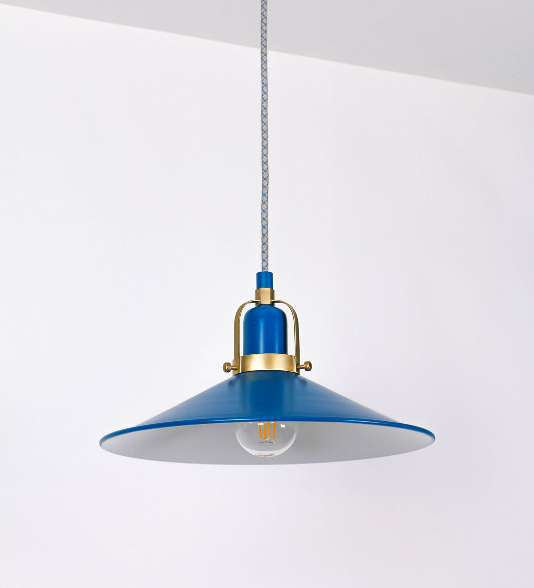 Suspensão de teto que combina um design clássico com cores modernas. A suspensão é fixa ao teto através de uma base circular azul vintage, de onde desce um cabo revestido têxtil com um padrão de linhas azuis e beges entrelaçadas, que sustenta o chapéu principal. O chapéu, azul vintage, tem um formato cónico invertido, com uma borda ligeiramente levantada. O interior do chapéu é branco. No topo do chapéu, há um suporte circular em latão escovado, com três pequenos braços que conecta o chapéu ao cabo.