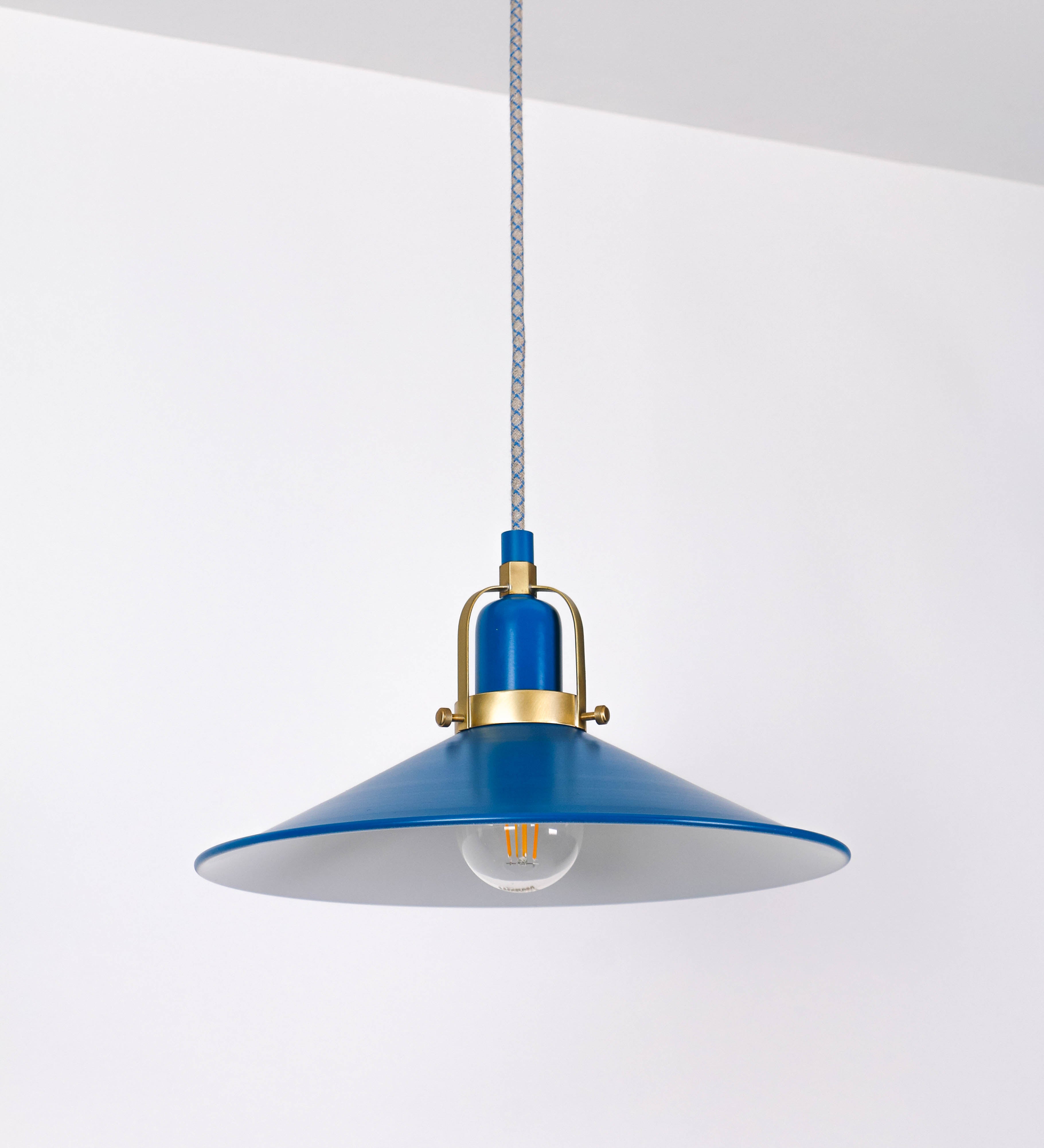 Suspensão de teto que combina um design clássico com cores modernas. A suspensão é fixa ao teto através de uma base circular azul vintage, de onde desce um cabo revestido têxtil com um padrão de linhas azuis e beges entrelaçadas, que sustenta o chapéu principal. O chapéu, azul vintage, tem um formato cónico invertido, com uma borda ligeiramente levantada. O interior do chapéu é branco. No topo do chapéu, há um suporte circular em latão escovado, com três pequenos braços que conecta o chapéu ao cabo.