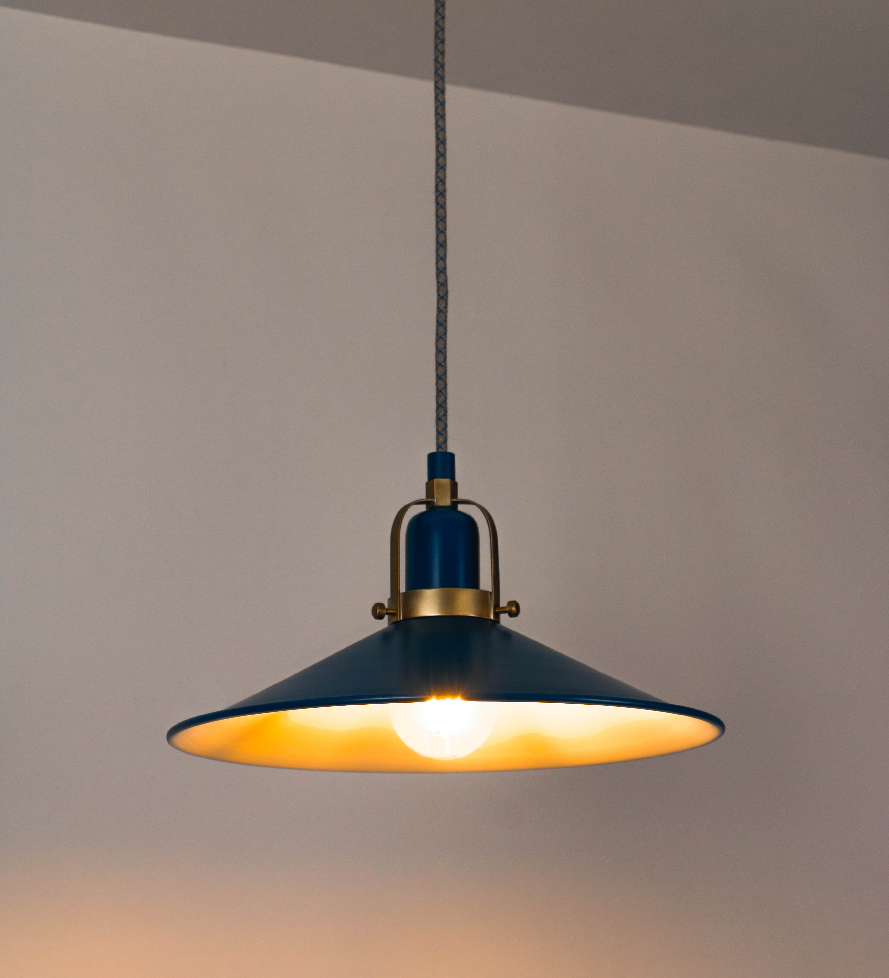 Suspensão de teto que combina um design clássico com cores modernas. A suspensão é fixa ao teto através de uma base circular azul vintage, de onde desce um cabo revestido têxtil com um padrão de linhas azuis e beges entrelaçadas, que sustenta o chapéu principal. O chapéu, azul vintage, tem um formato cónico invertido, com uma borda ligeiramente levantada. O interior do chapéu é branco. No topo do chapéu, há um suporte circular em latão escovado, com três pequenos braços que conecta o chapéu ao cabo.