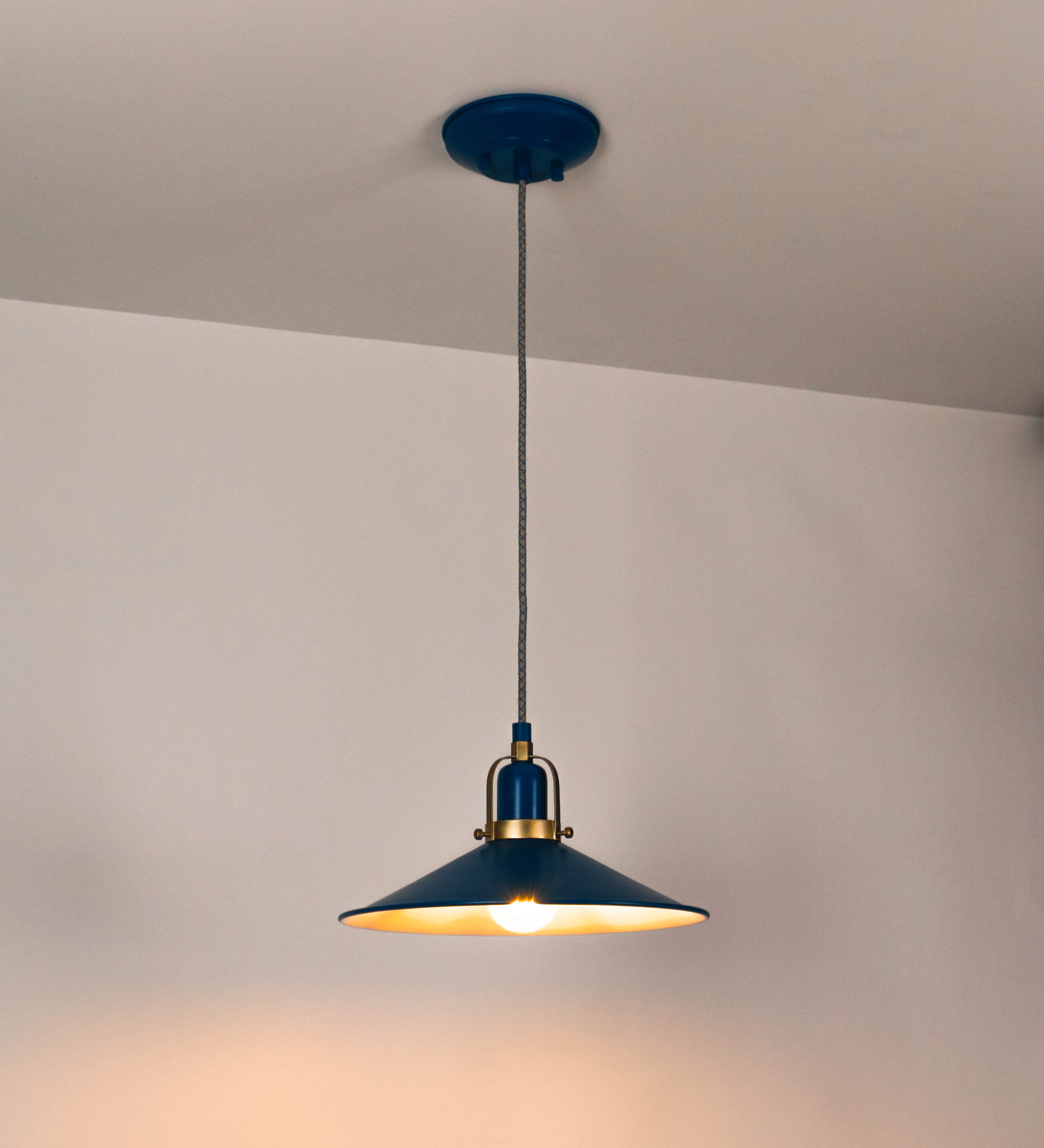 Suspensão de teto que combina um design clássico com cores modernas. A suspensão é fixa ao teto através de uma base circular azul vintage, de onde desce um cabo revestido têxtil com um padrão de linhas azuis e beges entrelaçadas, que sustenta o chapéu principal. O chapéu, azul vintage, tem um formato cónico invertido, com uma borda ligeiramente levantada. O interior do chapéu é branco. No topo do chapéu, há um suporte circular em latão escovado, com três pequenos braços que conecta o chapéu ao cabo.