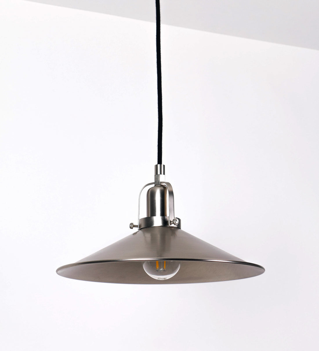 Suspensão de teto que combina um design clássico com um acabamento moderno e de estilo industrial. A suspensão, toda em níquel escovado, é fixa ao teto através de uma base circular de onde desce um cabo revestido têxtil preto, que sustenta o chapéu principal. O chapéu tem um formato cónico invertido, com uma borda ligeiramente levantada. No topo do chapéu, há um suporte circular com três pequenos braços que conecta o chapéu ao cabo.
