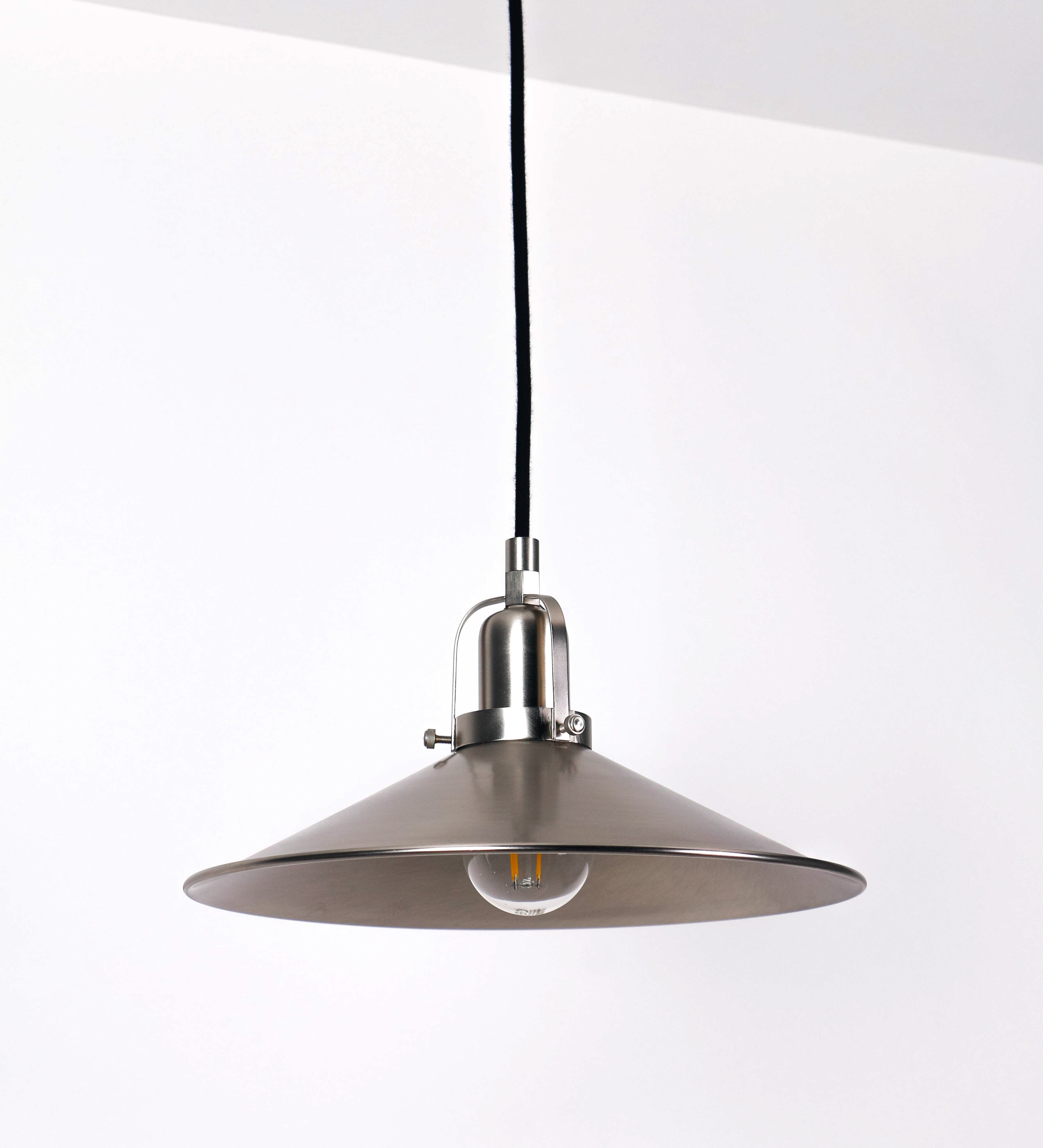 Suspensão de teto que combina um design clássico com um acabamento moderno e de estilo industrial. A suspensão, toda em níquel escovado, é fixa ao teto através de uma base circular de onde desce um cabo revestido têxtil preto, que sustenta o chapéu principal. O chapéu tem um formato cónico invertido, com uma borda ligeiramente levantada. No topo do chapéu, há um suporte circular com três pequenos braços que conecta o chapéu ao cabo.
