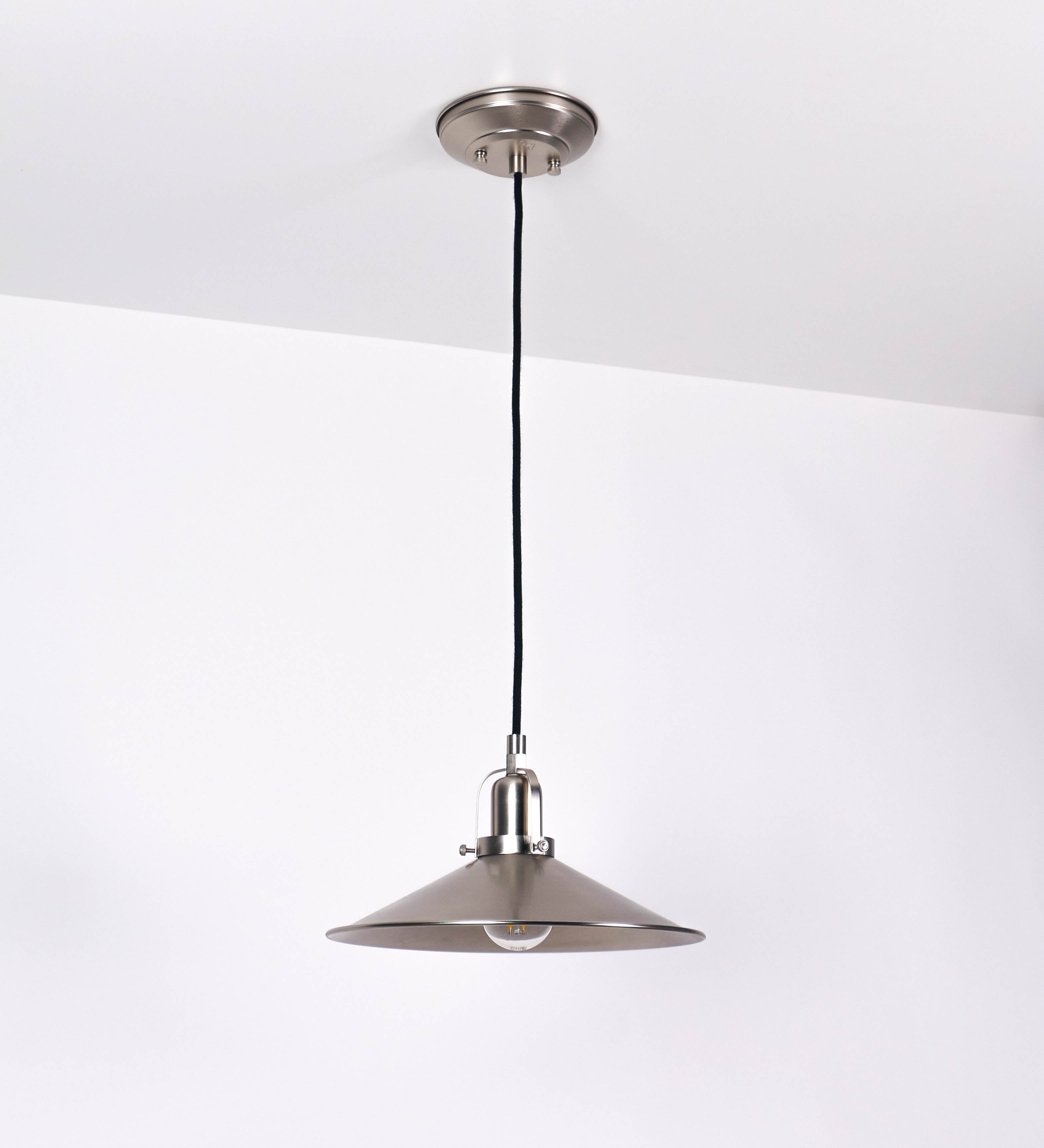 Suspensão de teto que combina um design clássico com um acabamento moderno e de estilo industrial. A suspensão, toda em níquel escovado, é fixa ao teto através de uma base circular de onde desce um cabo revestido têxtil preto, que sustenta o chapéu principal. O chapéu tem um formato cónico invertido, com uma borda ligeiramente levantada. No topo do chapéu, há um suporte circular com três pequenos braços que conecta o chapéu ao cabo.