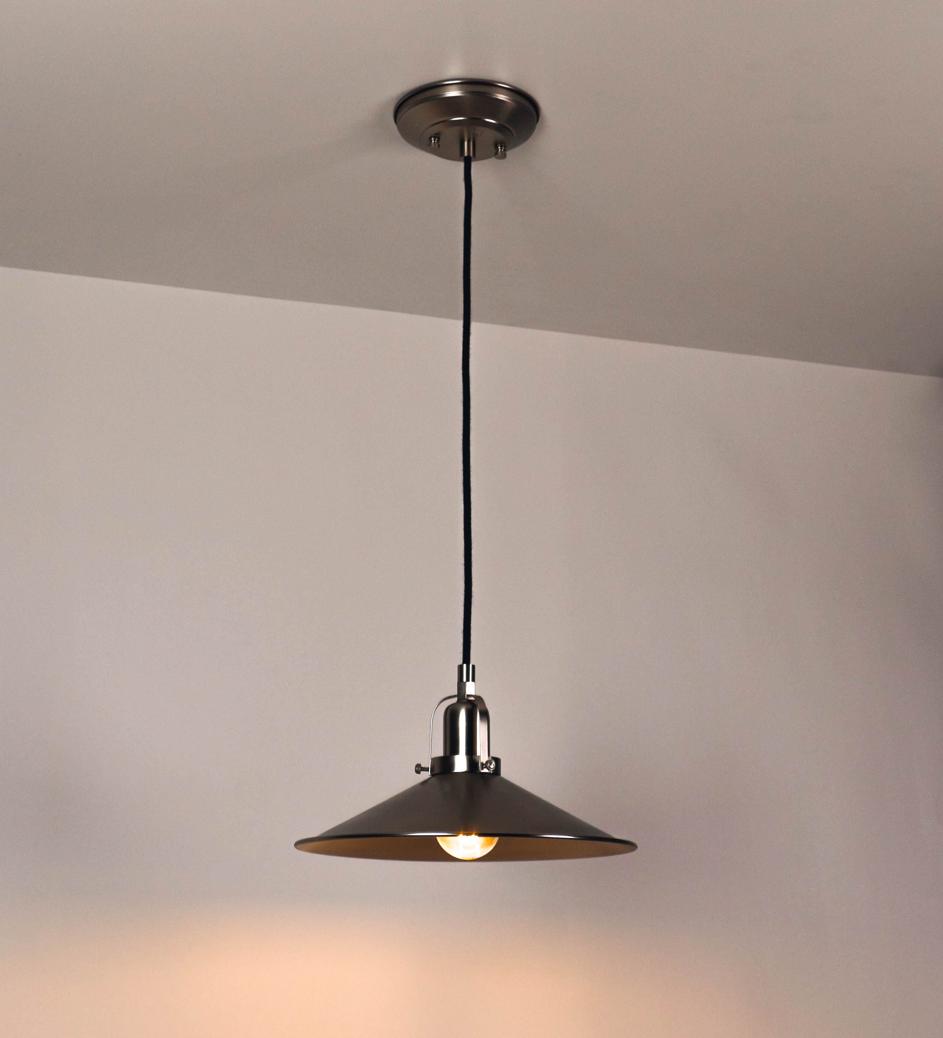 Suspensão de teto que combina um design clássico com um acabamento moderno e de estilo industrial. A suspensão, toda em níquel escovado, é fixa ao teto através de uma base circular de onde desce um cabo revestido têxtil preto, que sustenta o chapéu principal. O chapéu tem um formato cónico invertido, com uma borda ligeiramente levantada. No topo do chapéu, há um suporte circular com três pequenos braços que conecta o chapéu ao cabo.