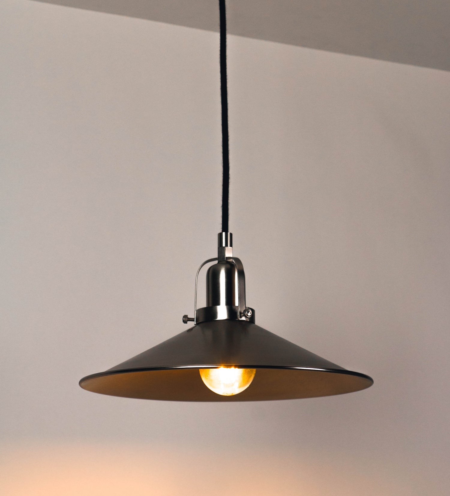 Suspensão de teto que combina um design clássico com um acabamento moderno e de estilo industrial. A suspensão, toda em níquel escovado, é fixa ao teto através de uma base circular de onde desce um cabo revestido têxtil preto, que sustenta o chapéu principal. O chapéu tem um formato cónico invertido, com uma borda ligeiramente levantada. No topo do chapéu, há um suporte circular com três pequenos braços que conecta o chapéu ao cabo.