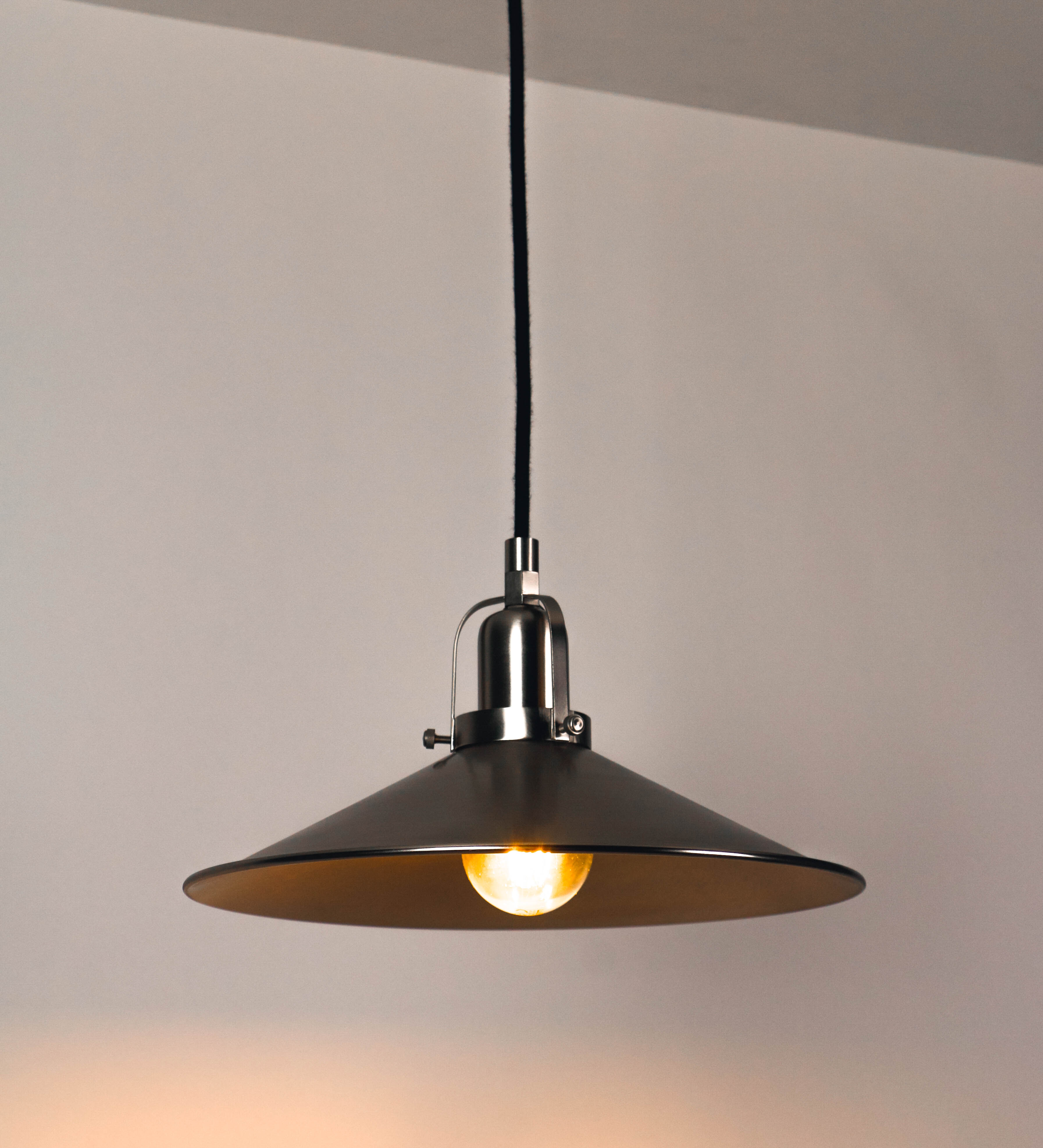 Suspensão de teto que combina um design clássico com um acabamento moderno e de estilo industrial. A suspensão, toda em níquel escovado, é fixa ao teto através de uma base circular de onde desce um cabo revestido têxtil preto, que sustenta o chapéu principal. O chapéu tem um formato cónico invertido, com uma borda ligeiramente levantada. No topo do chapéu, há um suporte circular com três pequenos braços que conecta o chapéu ao cabo.