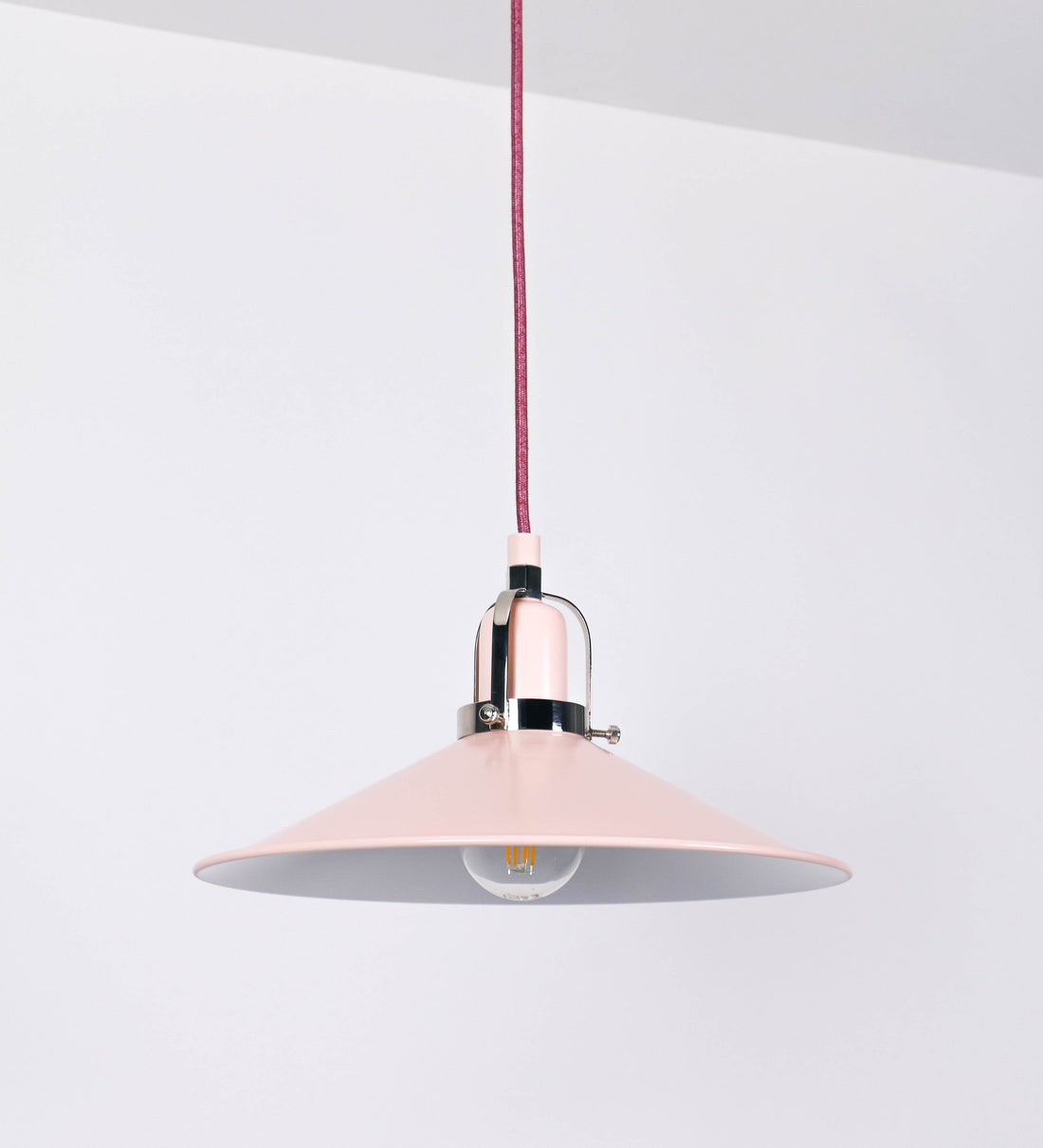 Suspensão de teto que combina um design clássico com cores modernas. A suspensão é fixa ao teto através de uma base circular rosa clara, de onde desce um cabo revestido têxtil rosa velho, que sustenta o chapéu principal. O chapéu, rosa claro, tem um formato cónico invertido, com uma borda ligeiramente levantada. O interior do chapéu é branco. No topo do chapéu, há um suporte circular em níquel polido, com três pequenos braços que conecta o chapéu ao cabo.