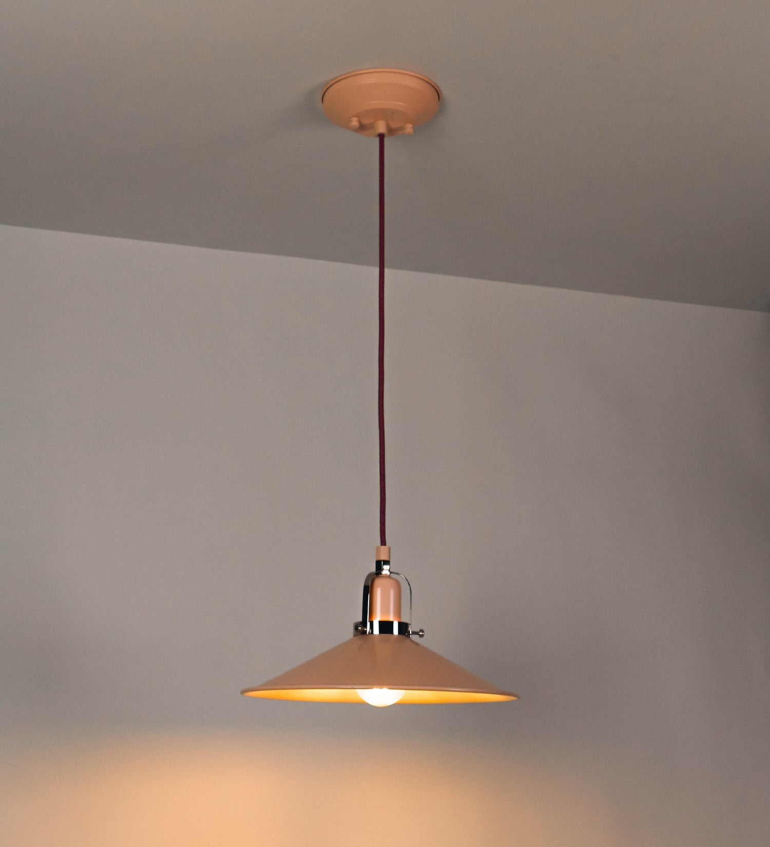 Suspensão de teto que combina um design clássico com cores modernas. A suspensão é fixa ao teto através de uma base circular rosa clara, de onde desce um cabo revestido têxtil rosa velho, que sustenta o chapéu principal. O chapéu, rosa claro, tem um formato cónico invertido, com uma borda ligeiramente levantada. O interior do chapéu é branco. No topo do chapéu, há um suporte circular em níquel polido, com três pequenos braços que conecta o chapéu ao cabo.