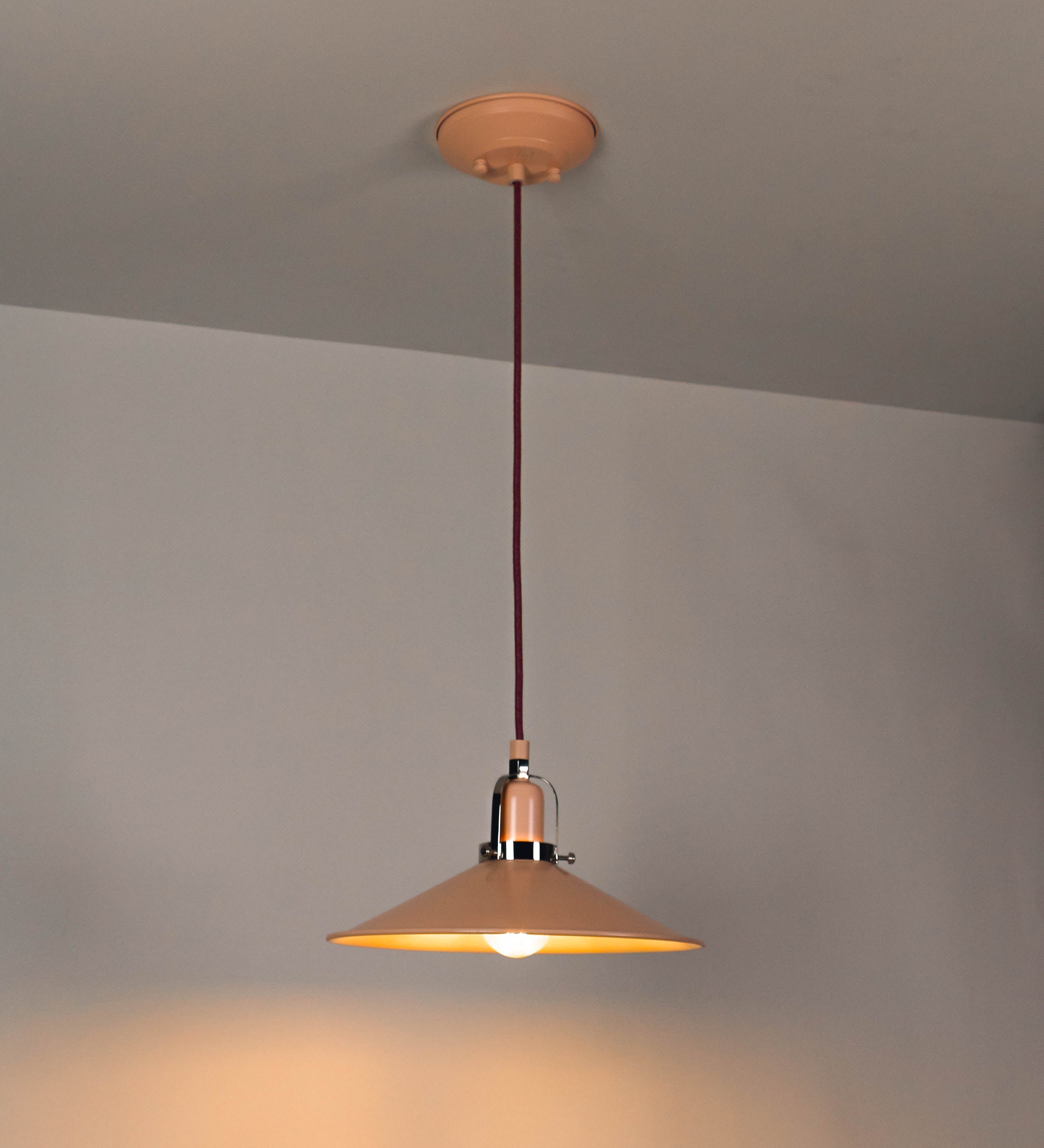 Suspensão de teto que combina um design clássico com cores modernas. A suspensão é fixa ao teto através de uma base circular rosa clara, de onde desce um cabo revestido têxtil rosa velho, que sustenta o chapéu principal. O chapéu, rosa claro, tem um formato cónico invertido, com uma borda ligeiramente levantada. O interior do chapéu é branco. No topo do chapéu, há um suporte circular em níquel polido, com três pequenos braços que conecta o chapéu ao cabo.