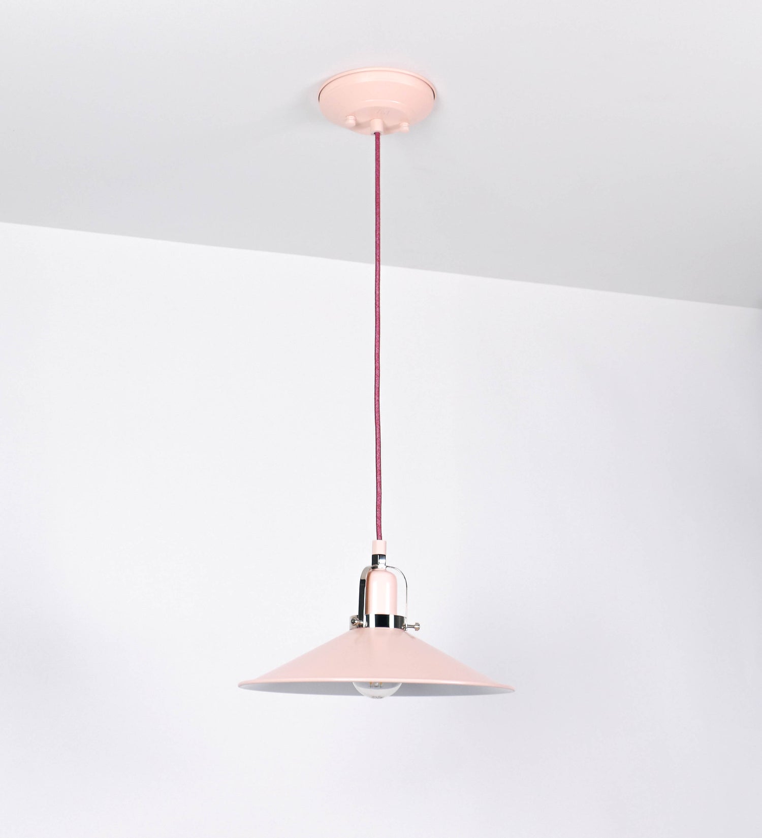 Suspensão de teto que combina um design clássico com cores modernas. A suspensão é fixa ao teto através de uma base circular rosa clara, de onde desce um cabo revestido têxtil rosa velho, que sustenta o chapéu principal. O chapéu, rosa claro, tem um formato cónico invertido, com uma borda ligeiramente levantada. O interior do chapéu é branco. No topo do chapéu, há um suporte circular em níquel polido, com três pequenos braços que conecta o chapéu ao cabo.