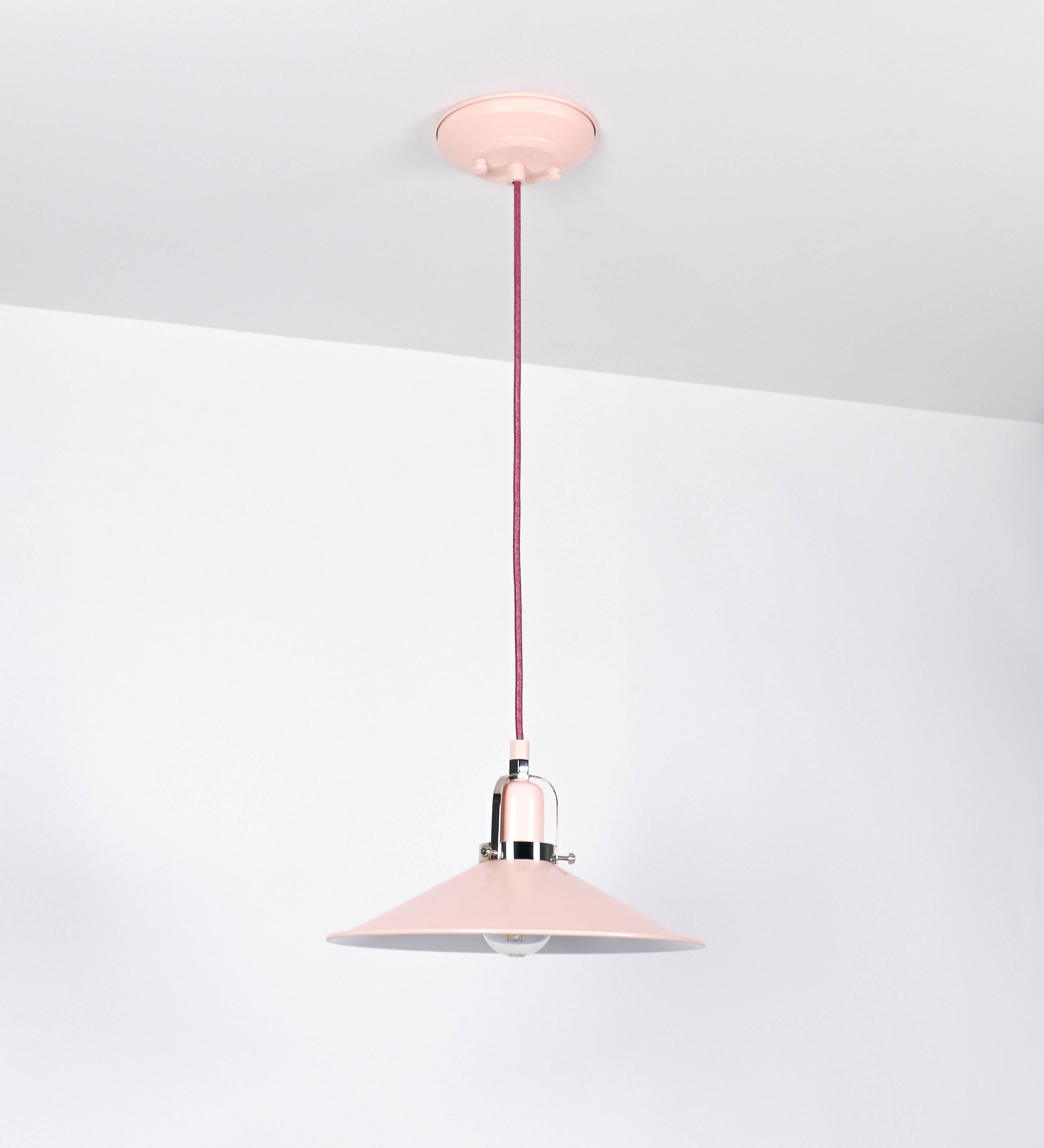 Suspensão de teto que combina um design clássico com cores modernas. A suspensão é fixa ao teto através de uma base circular rosa clara, de onde desce um cabo revestido têxtil rosa velho, que sustenta o chapéu principal. O chapéu, rosa claro, tem um formato cónico invertido, com uma borda ligeiramente levantada. O interior do chapéu é branco. No topo do chapéu, há um suporte circular em níquel polido, com três pequenos braços que conecta o chapéu ao cabo.