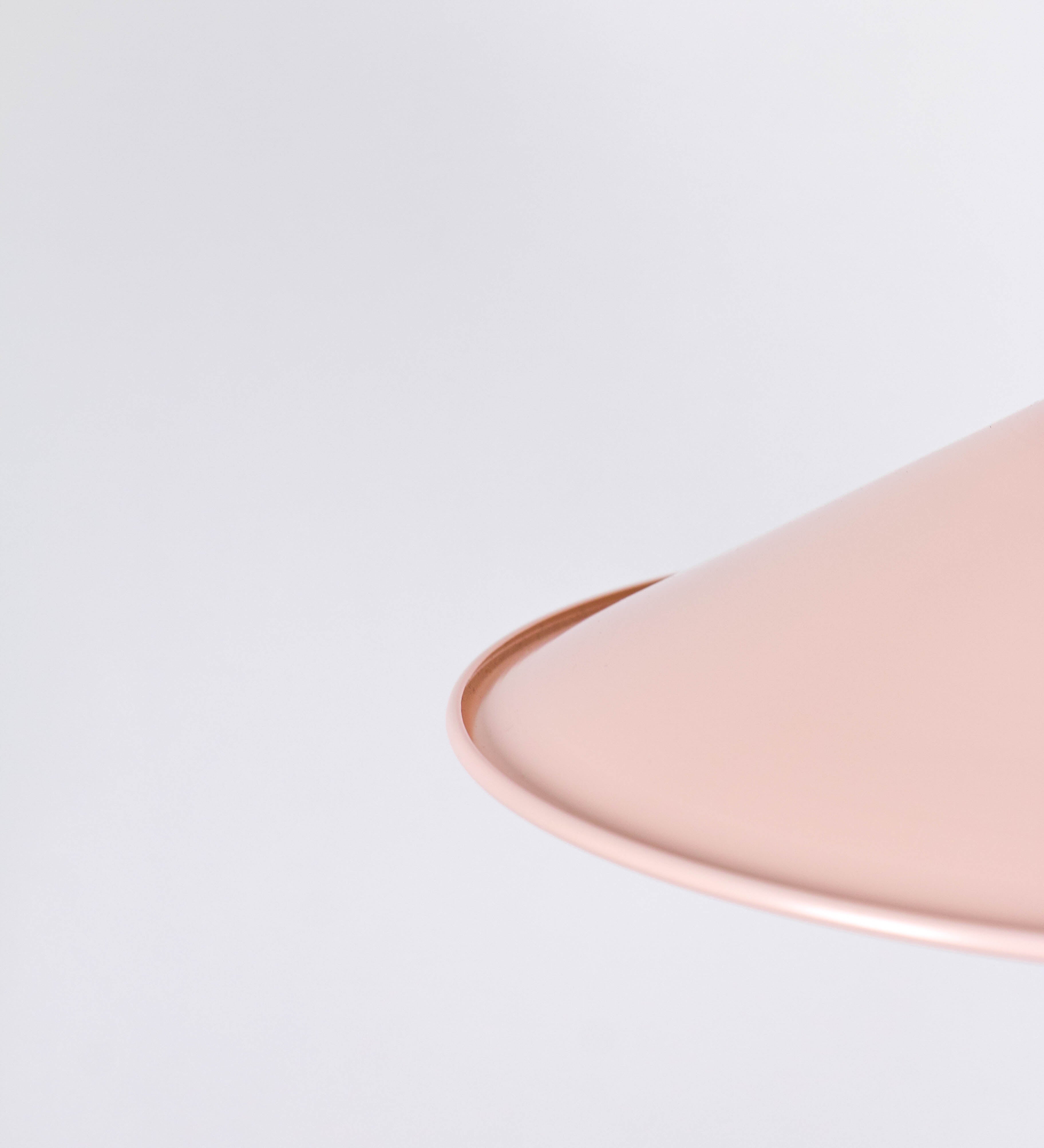 Suspensão de teto que combina um design clássico com cores modernas. A suspensão é fixa ao teto através de uma base circular rosa clara, de onde desce um cabo revestido têxtil rosa velho, que sustenta o chapéu principal. O chapéu, rosa claro, tem um formato cónico invertido, com uma borda ligeiramente levantada. O interior do chapéu é branco. No topo do chapéu, há um suporte circular em níquel polido, com três pequenos braços que conecta o chapéu ao cabo.
