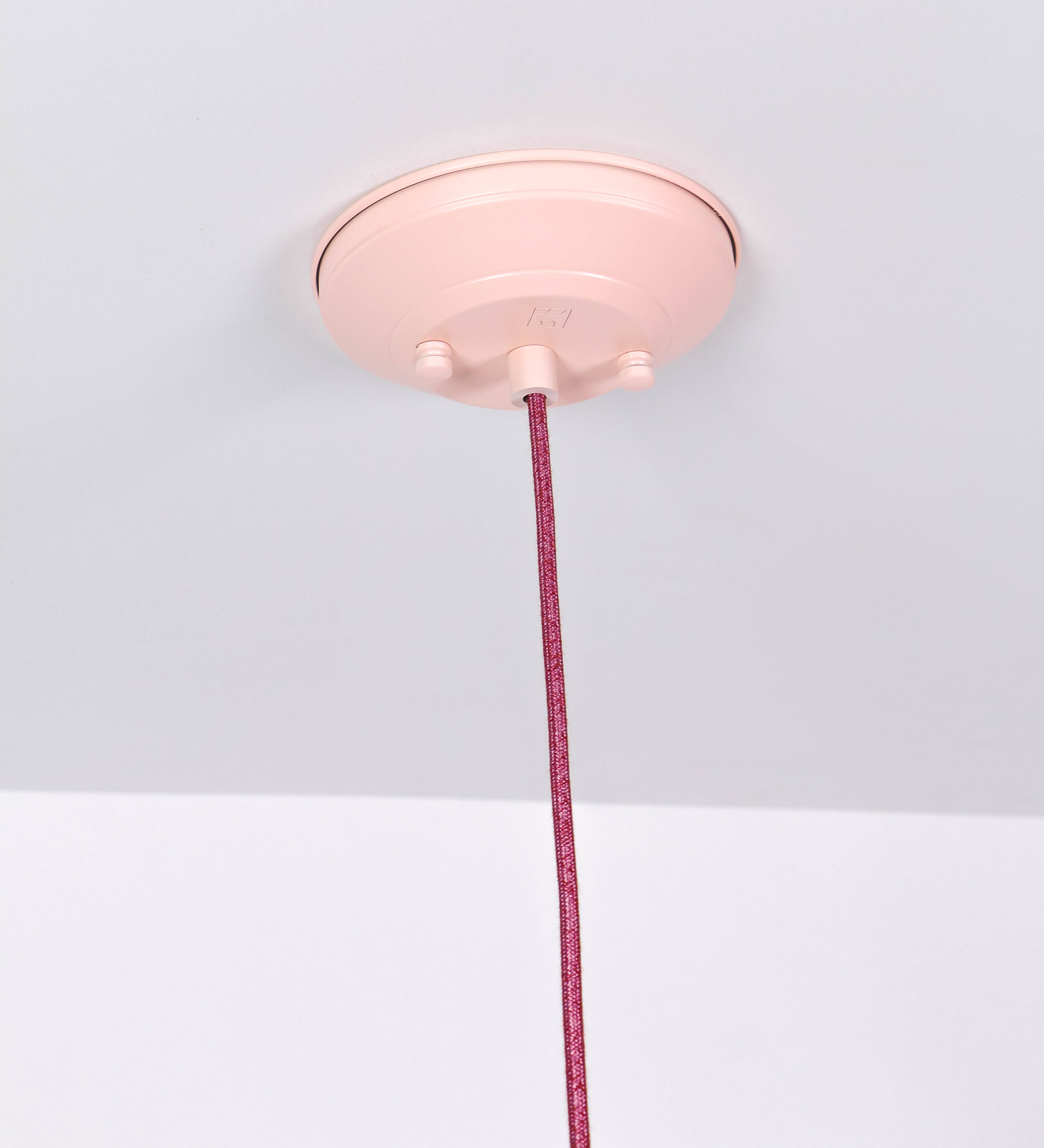 Suspensão de teto que combina um design clássico com cores modernas. A suspensão é fixa ao teto através de uma base circular rosa clara, de onde desce um cabo revestido têxtil rosa velho, que sustenta o chapéu principal. O chapéu, rosa claro, tem um formato cónico invertido, com uma borda ligeiramente levantada. O interior do chapéu é branco. No topo do chapéu, há um suporte circular em níquel polido, com três pequenos braços que conecta o chapéu ao cabo.