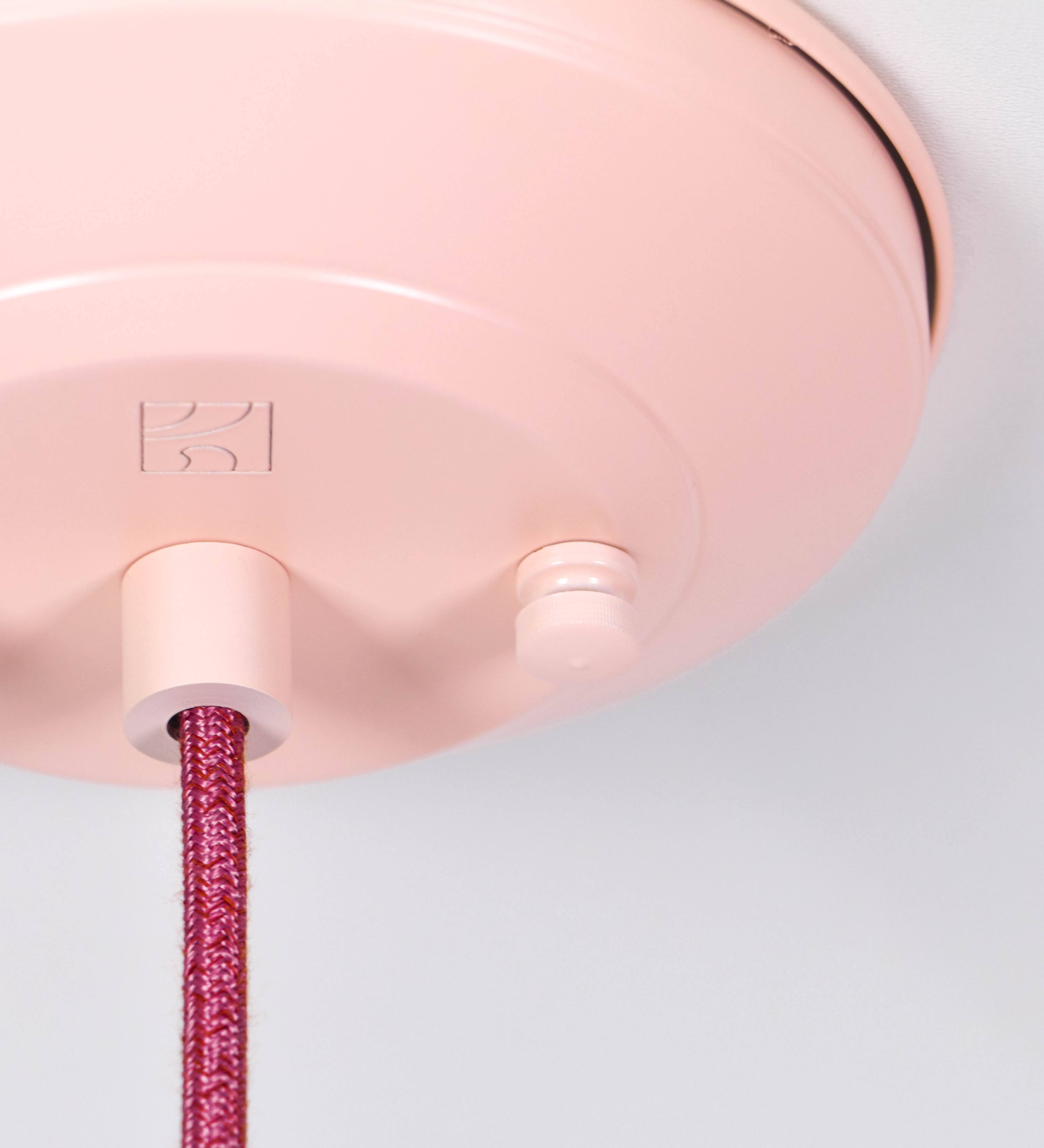Suspensão de teto que combina um design clássico com cores modernas. A suspensão é fixa ao teto através de uma base circular rosa clara, de onde desce um cabo revestido têxtil rosa velho, que sustenta o chapéu principal. O chapéu, rosa claro, tem um formato cónico invertido, com uma borda ligeiramente levantada. O interior do chapéu é branco. No topo do chapéu, há um suporte circular em níquel polido, com três pequenos braços que conecta o chapéu ao cabo.
