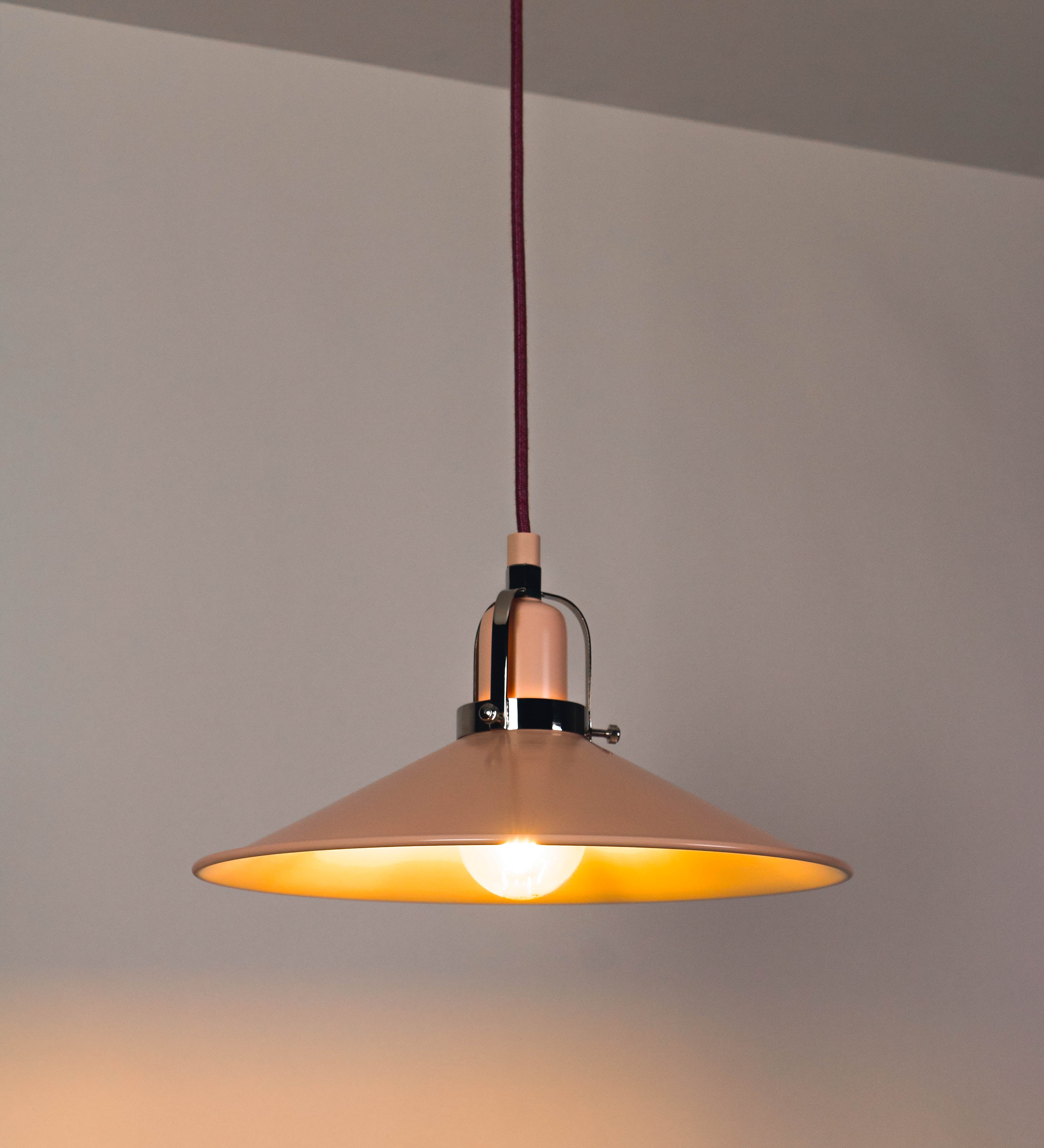 Suspensão de teto que combina um design clássico com cores modernas. A suspensão é fixa ao teto através de uma base circular rosa clara, de onde desce um cabo revestido têxtil rosa velho, que sustenta o chapéu principal. O chapéu, rosa claro, tem um formato cónico invertido, com uma borda ligeiramente levantada. O interior do chapéu é branco. No topo do chapéu, há um suporte circular em níquel polido, com três pequenos braços que conecta o chapéu ao cabo.