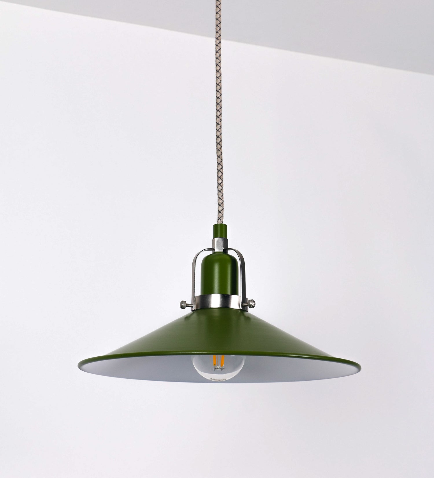 Suspensão de teto que combina um design clássico e cores modernas. A suspensão é fixa ao teto através de uma base circular verde couve-de-bruxelas, de onde desce um cabo revestido têxtil com um padrão de linhas castanhas e beges entrelaçadas, que sustenta o chapéu principal. O chapéu, verde, tem um formato cónico invertido, com uma borda ligeiramente levantada. O interior do chapéu é branco. No topo do chapéu, há um suporte circular em níquel escovado, com três pequenos braços que conecta o chapéu ao cabo.