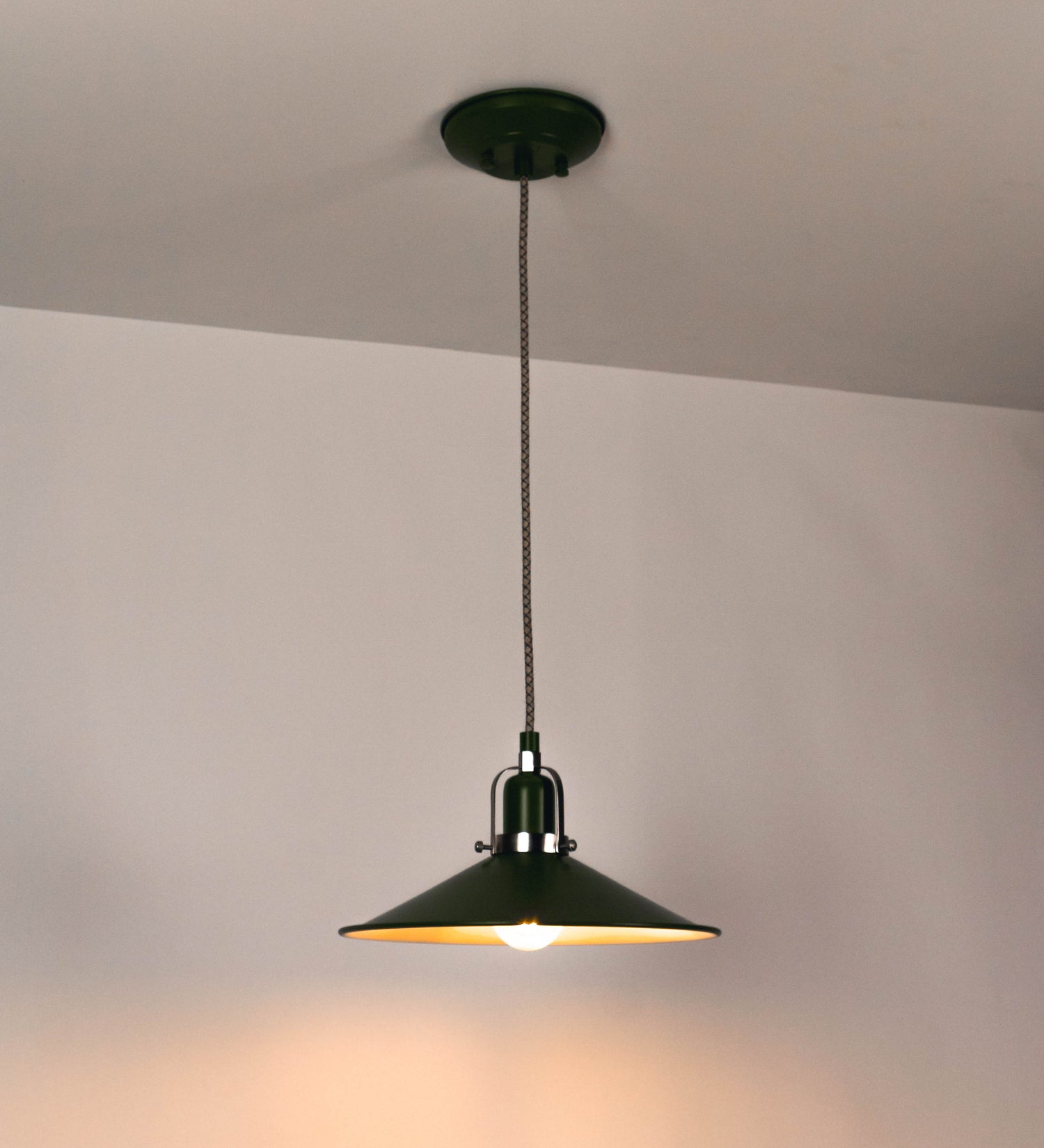 Suspensão de teto que combina um design clássico e cores modernas. A suspensão é fixa ao teto através de uma base circular verde couve-de-bruxelas, de onde desce um cabo revestido têxtil com um padrão de linhas castanhas e beges entrelaçadas, que sustenta o chapéu principal. O chapéu, verde, tem um formato cónico invertido, com uma borda ligeiramente levantada. O interior do chapéu é branco. No topo do chapéu, há um suporte circular em níquel escovado, com três pequenos braços que conecta o chapéu ao cabo.