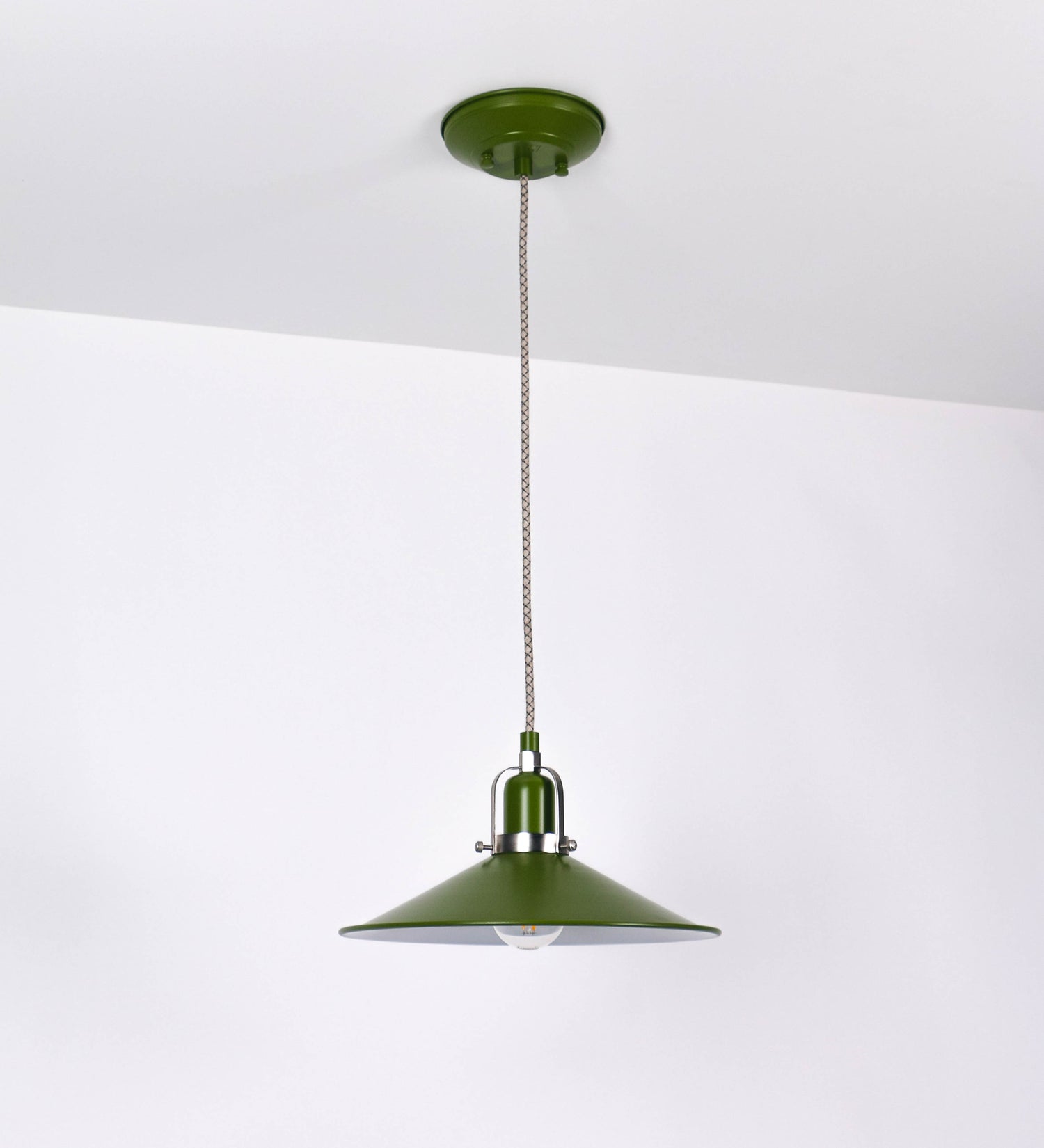 Suspensão de teto que combina um design clássico e cores modernas. A suspensão é fixa ao teto através de uma base circular verde couve-de-bruxelas, de onde desce um cabo revestido têxtil com um padrão de linhas castanhas e beges entrelaçadas, que sustenta o chapéu principal. O chapéu, verde, tem um formato cónico invertido, com uma borda ligeiramente levantada. O interior do chapéu é branco. No topo do chapéu, há um suporte circular em níquel escovado, com três pequenos braços que conecta o chapéu ao cabo.