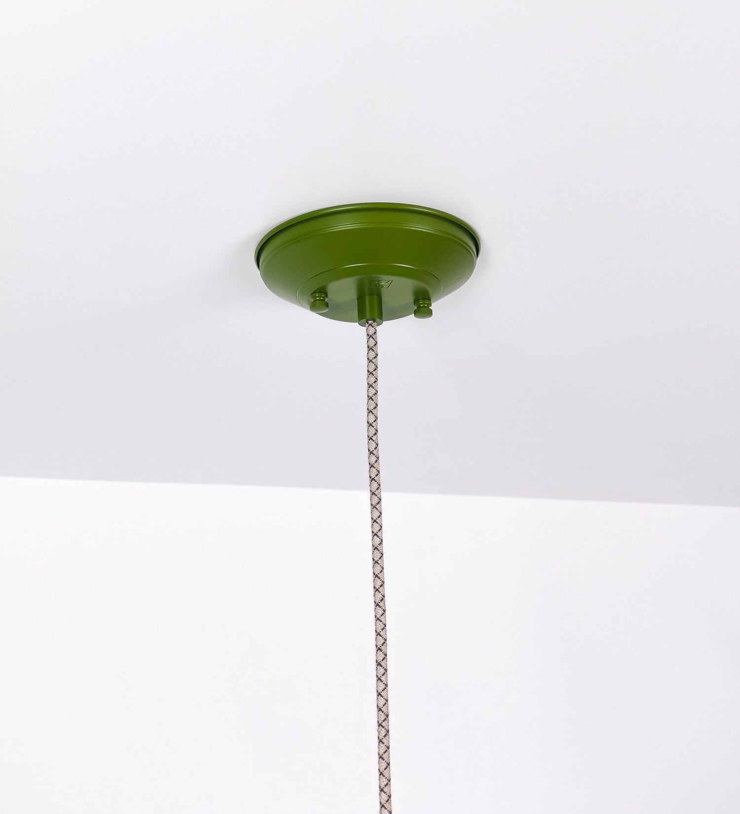 Suspensão de teto que combina um design clássico e cores modernas. A suspensão é fixa ao teto através de uma base circular verde couve-de-bruxelas, de onde desce um cabo revestido têxtil com um padrão de linhas castanhas e beges entrelaçadas, que sustenta o chapéu principal. O chapéu, verde, tem um formato cónico invertido, com uma borda ligeiramente levantada. O interior do chapéu é branco. No topo do chapéu, há um suporte circular em níquel escovado, com três pequenos braços que conecta o chapéu ao cabo.