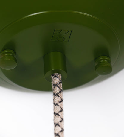 Suspensão de teto que combina um design clássico e cores modernas. A suspensão é fixa ao teto através de uma base circular verde couve-de-bruxelas, de onde desce um cabo revestido têxtil com um padrão de linhas castanhas e beges entrelaçadas, que sustenta o chapéu principal. O chapéu, verde, tem um formato cónico invertido, com uma borda ligeiramente levantada. O interior do chapéu é branco. No topo do chapéu, há um suporte circular em níquel escovado, com três pequenos braços que conecta o chapéu ao cabo.