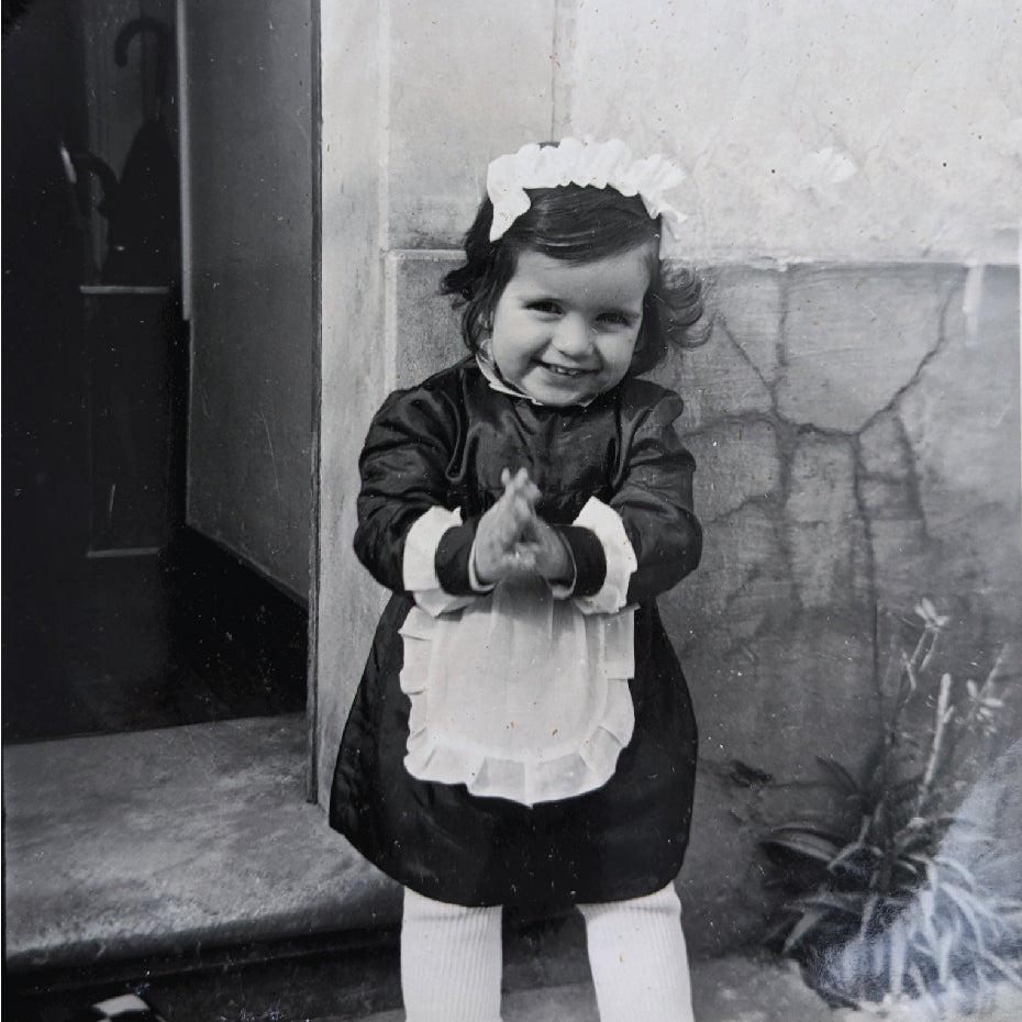 Fotografia antiga em preto e branco de uma criança, Ana Faria, filha do fundador da Lustrarte, sorrindo à porta de casa nos primeiros anos da marca.